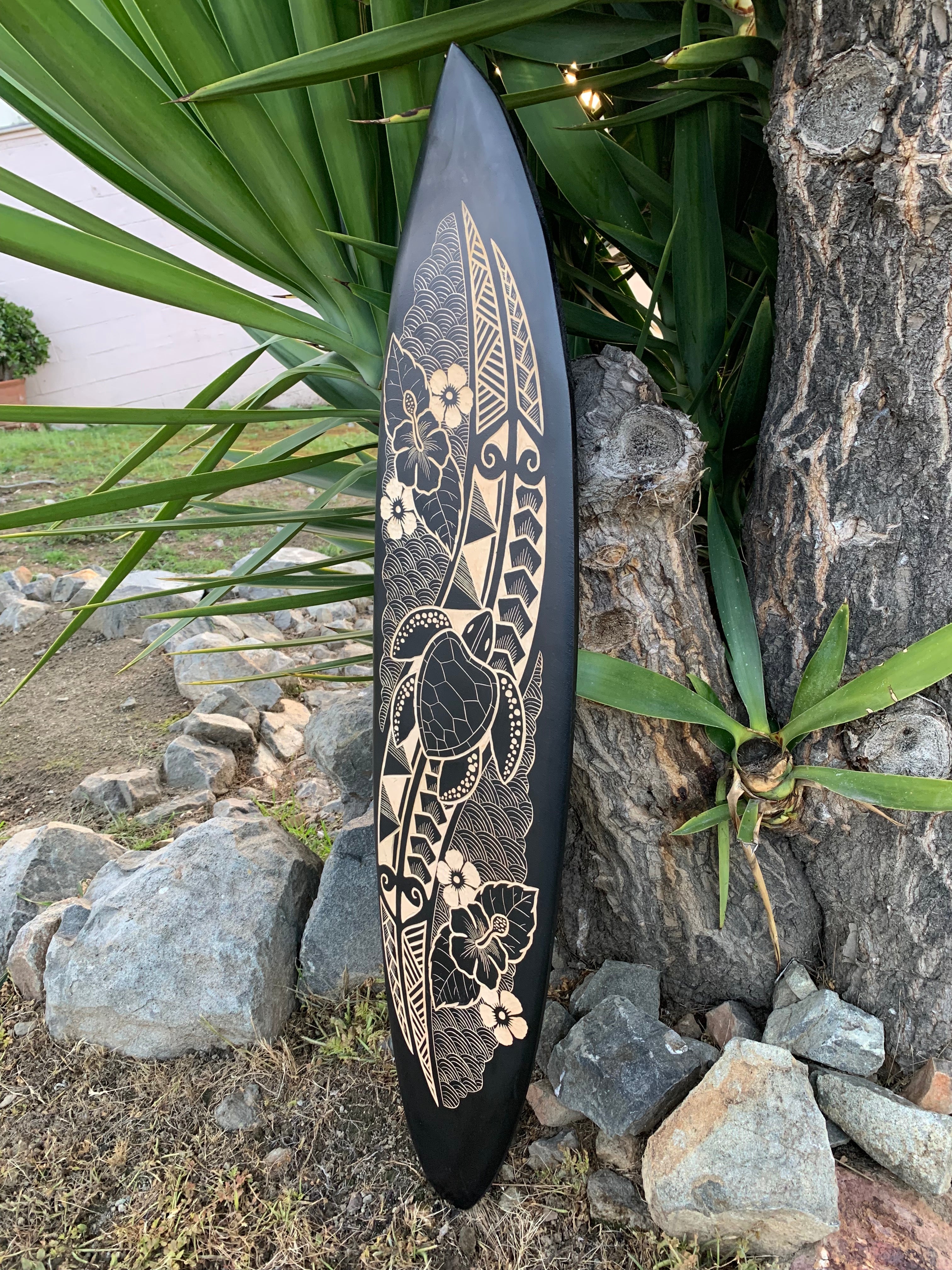 black decorative surfboard with beige tropical flower and leaf engravings leaning against a tree