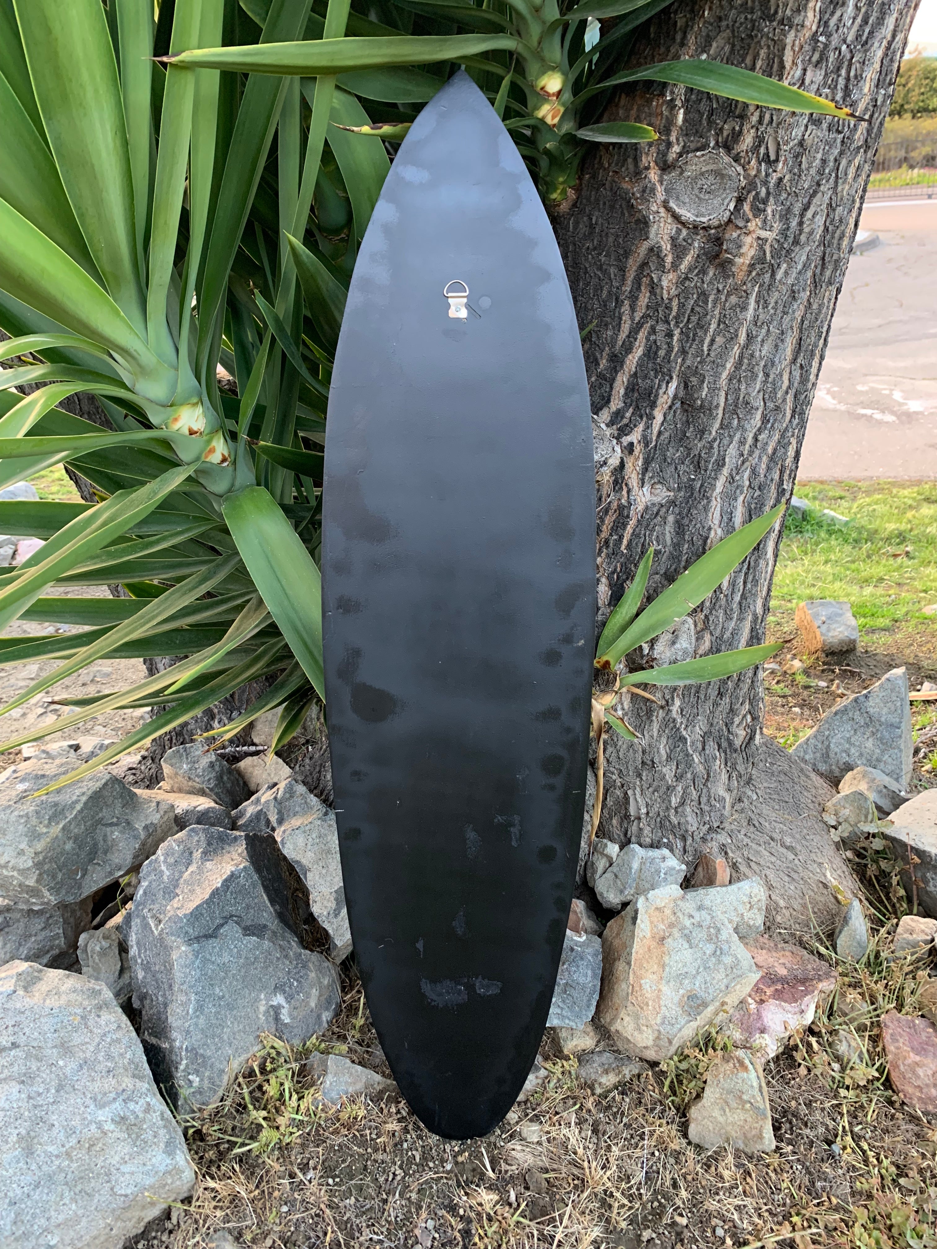 Black surfboard leaning against a tree amid plants and rocks.
