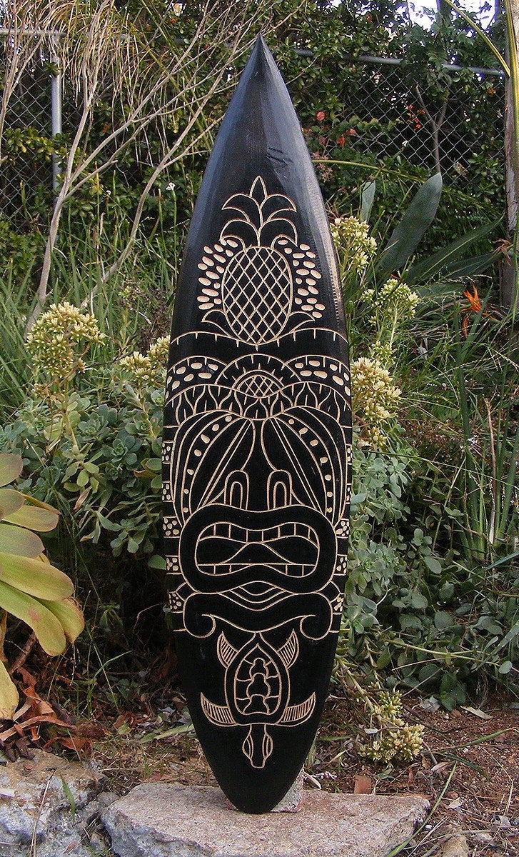 Black surfboard with beige tiki carving featuring pineapple motif, standing upright in a garden.
