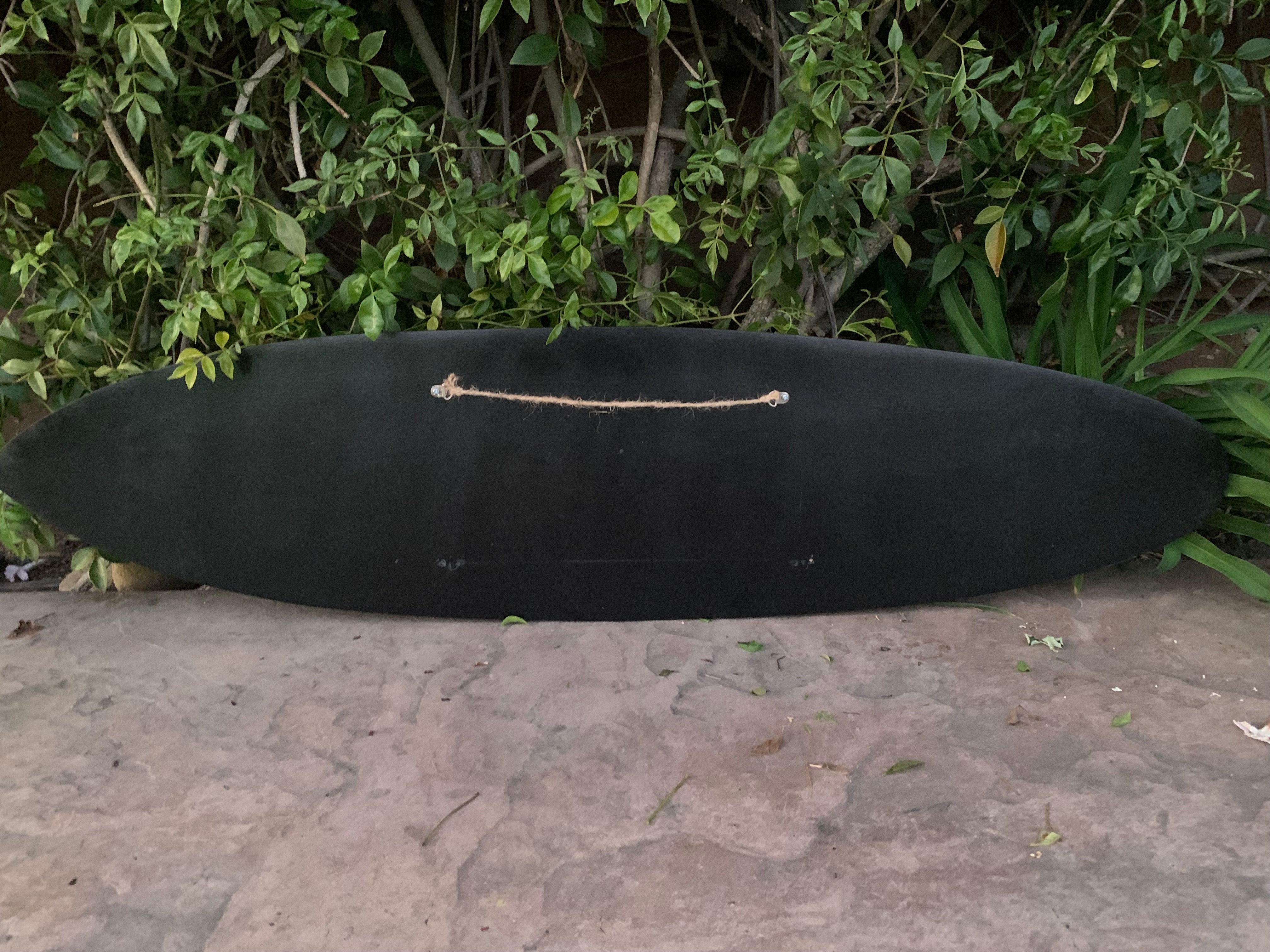 Black surfboard with a rope handle resting on a concrete surface beside green bushes.
