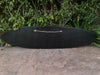 Black surfboard with a rope handle resting on a concrete surface beside green bushes.