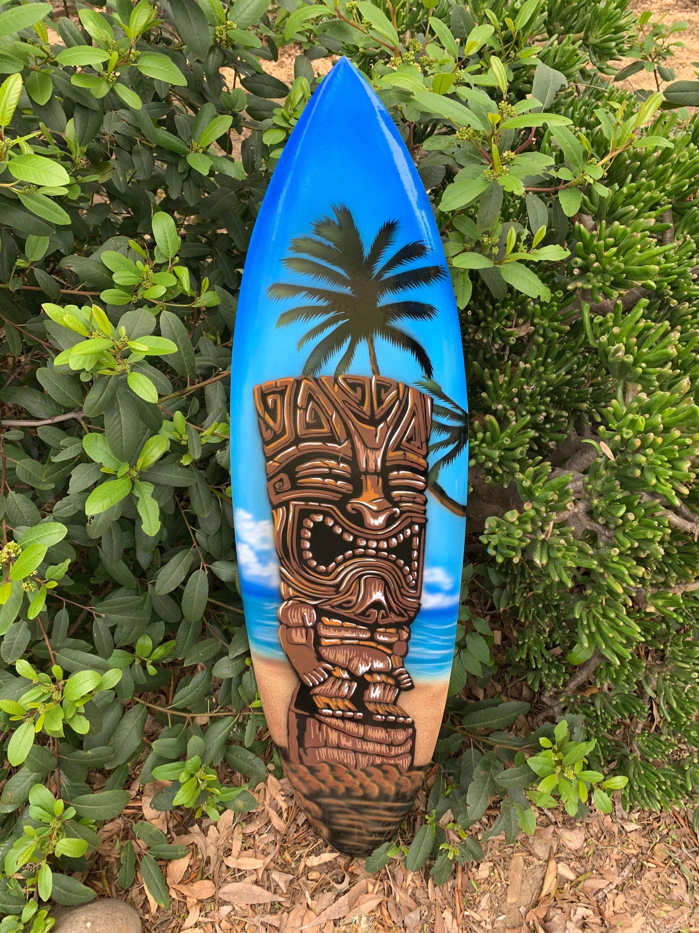 blue surfboard featuring a tiki face and a palm tree on a tropical beach