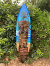 Blue surfboard with Polynesian tiki carving and palm tree design leaning against tropical greenery.