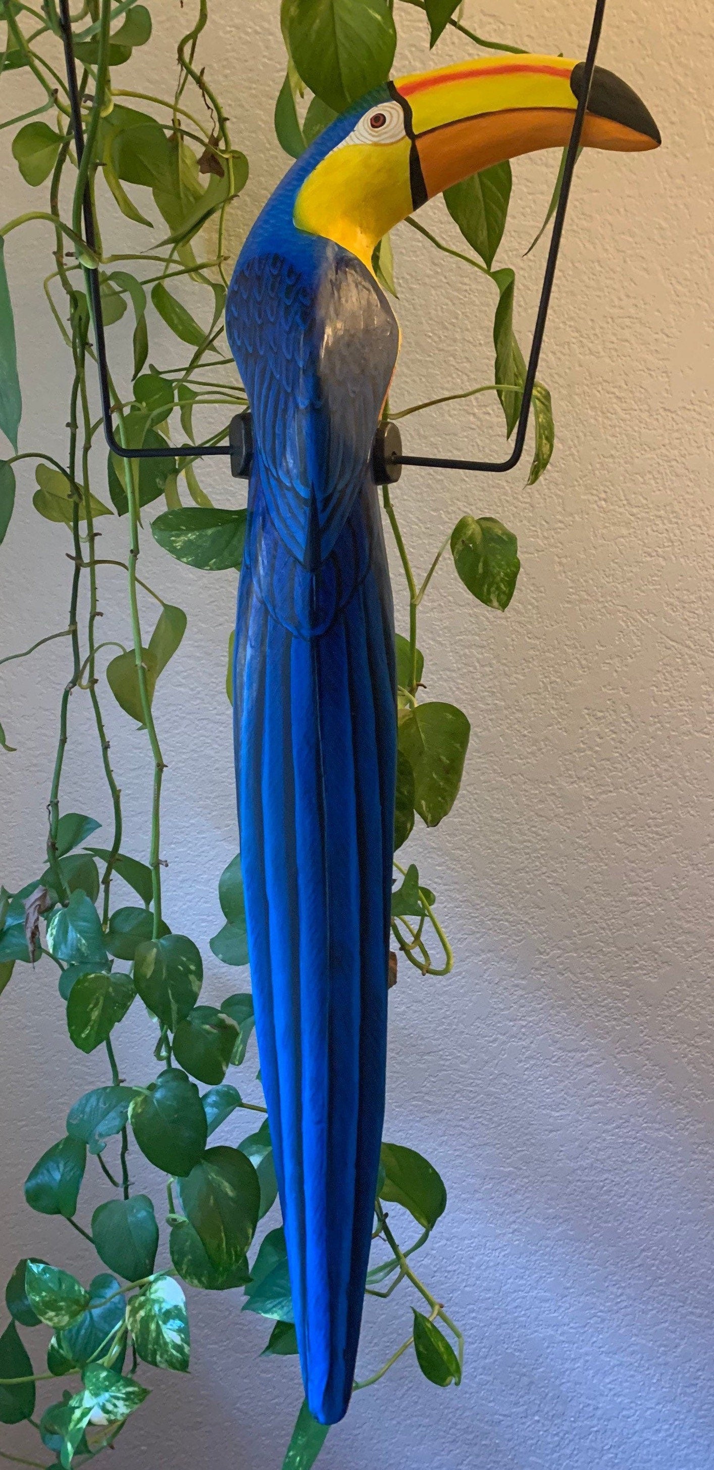 Decorative blue-and-yellow macaw sculpture with a long tail perched on a metal stand beside green houseplants.