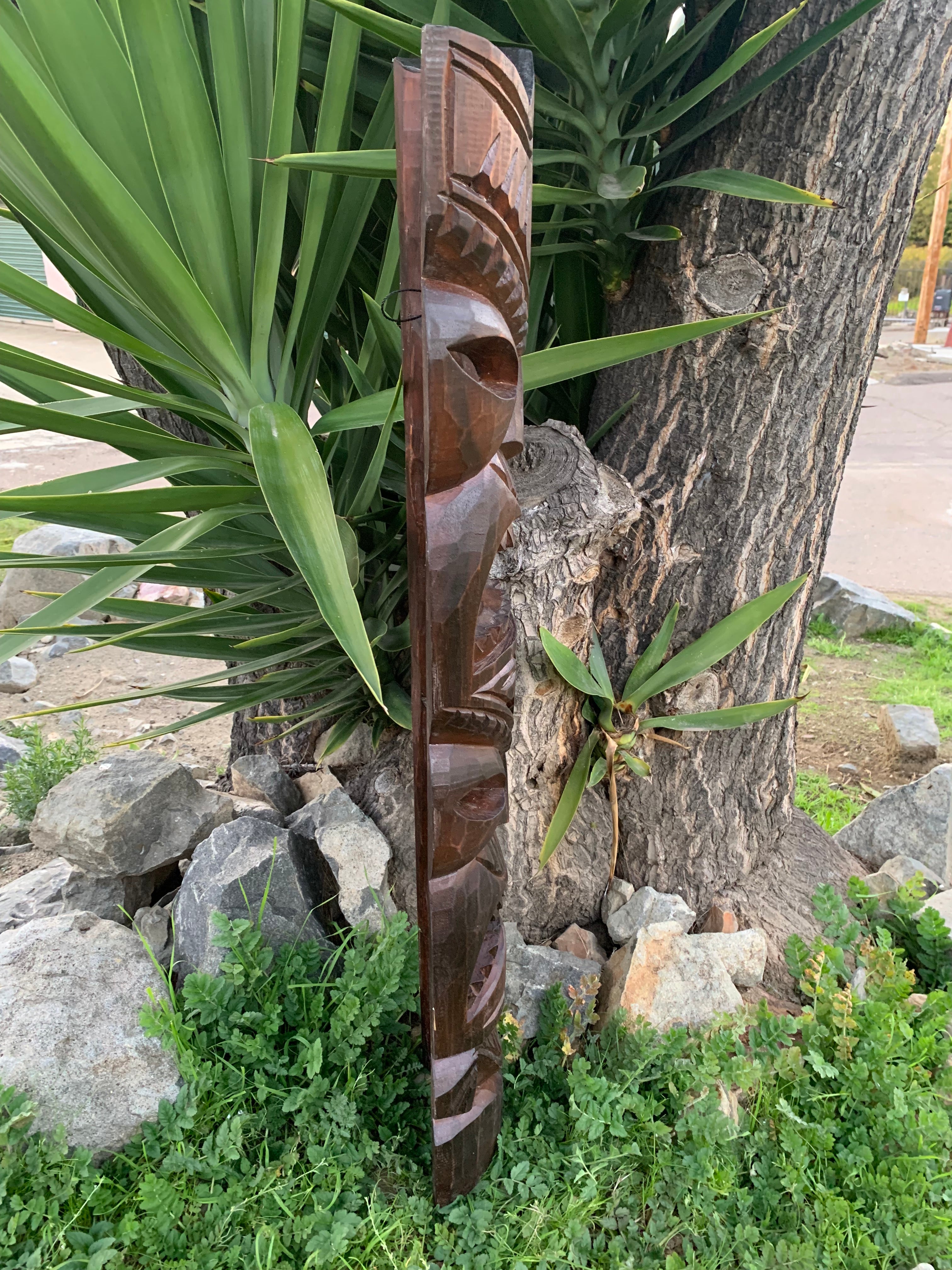 brown carved wooden totem sculpture leaning against a tree beside green Yucca plants and rocks