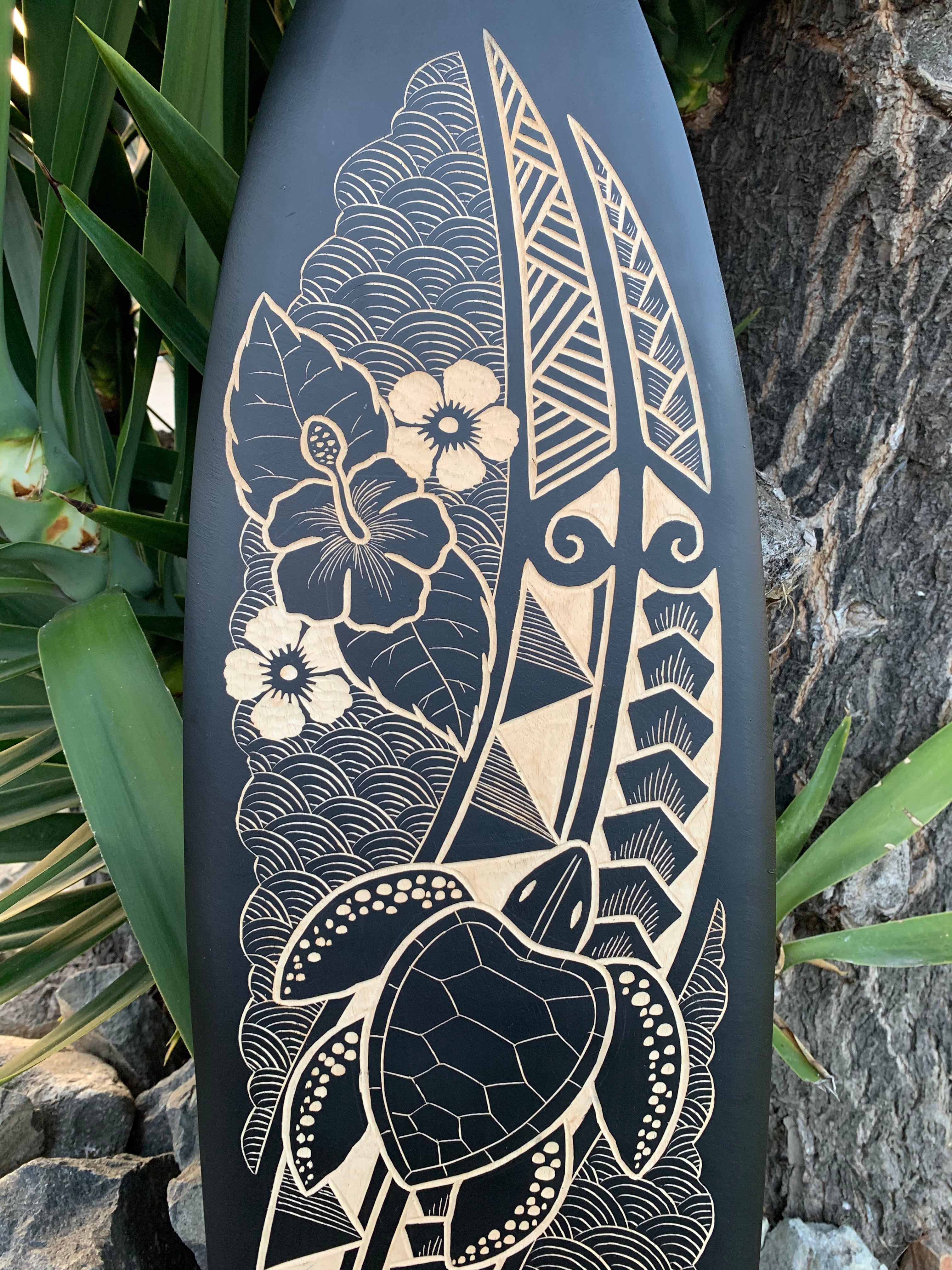 Carved surfboard with a turtle, hibiscus flower, and wave patterns in beige on a dark background