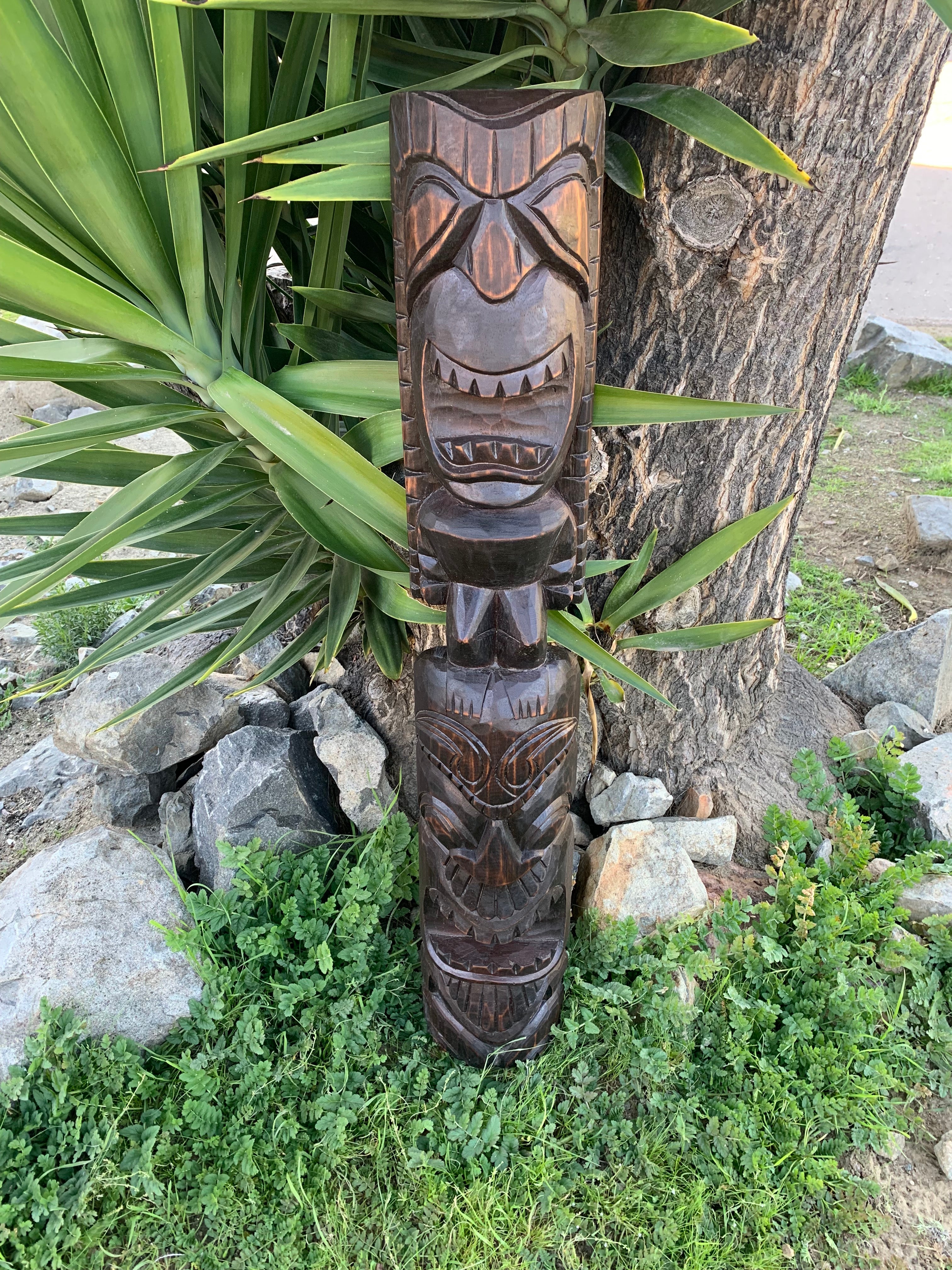 Dark wooden tiki totem pole carved with multiple faces outdoors beside a tree and greenery