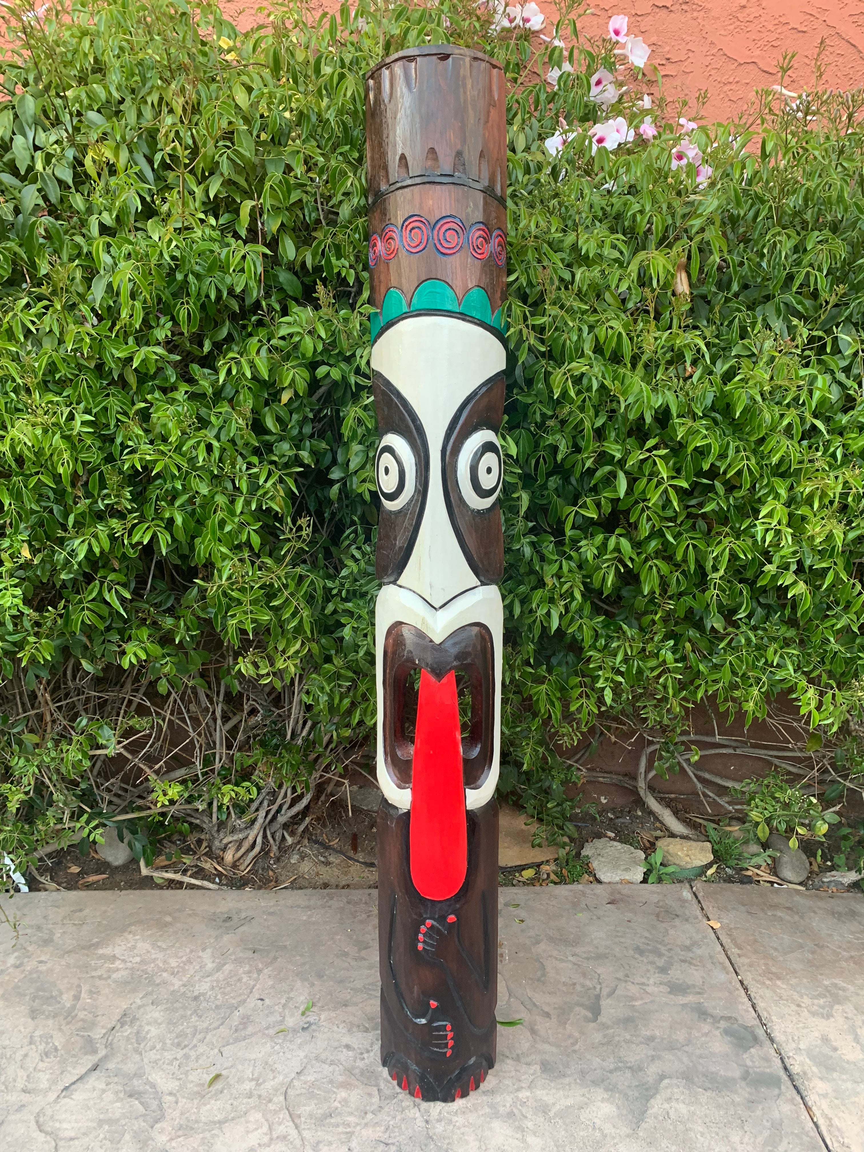 Carved tiki totem pole with a wide white face, black eye outlines, and a red tongue, outdoors