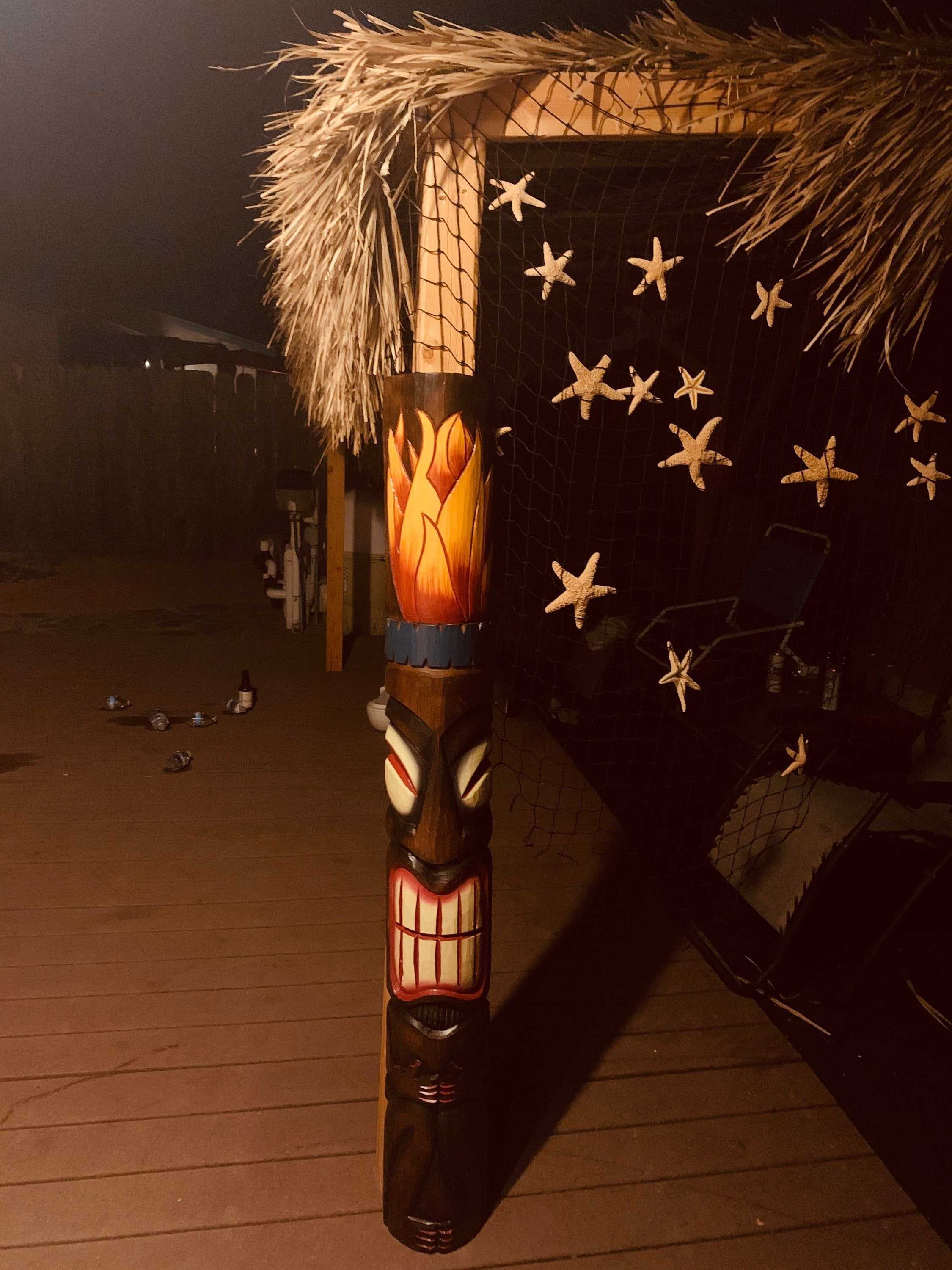 Carved tiki totem pole on a beach deck with thatched roof and starfish netting at night.