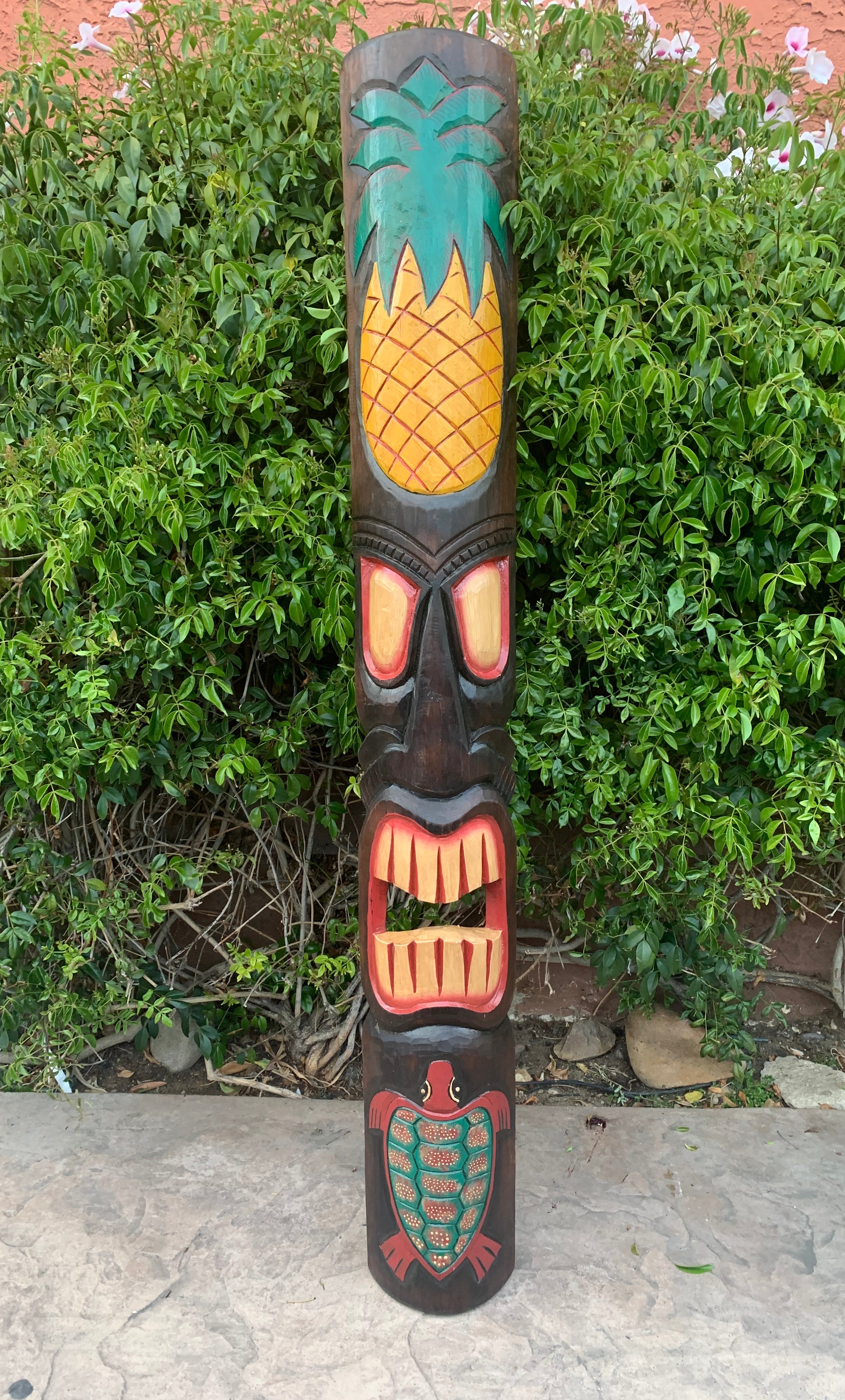 Colorful wooden totem pole with pineapple top, bold eyes, open mouth, and turtle base.