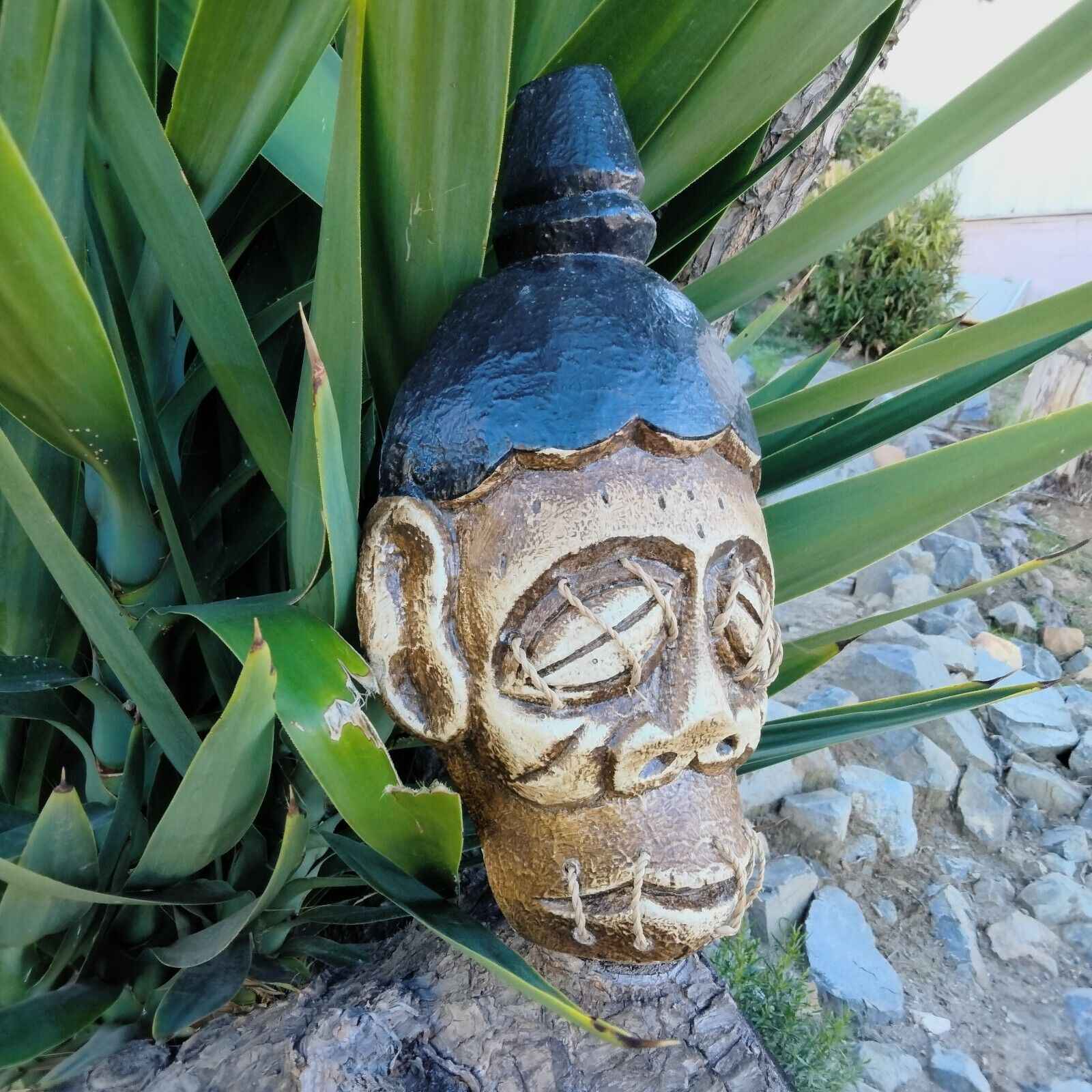 carved tribal mask head with blue hat among green plant leaves in garden
