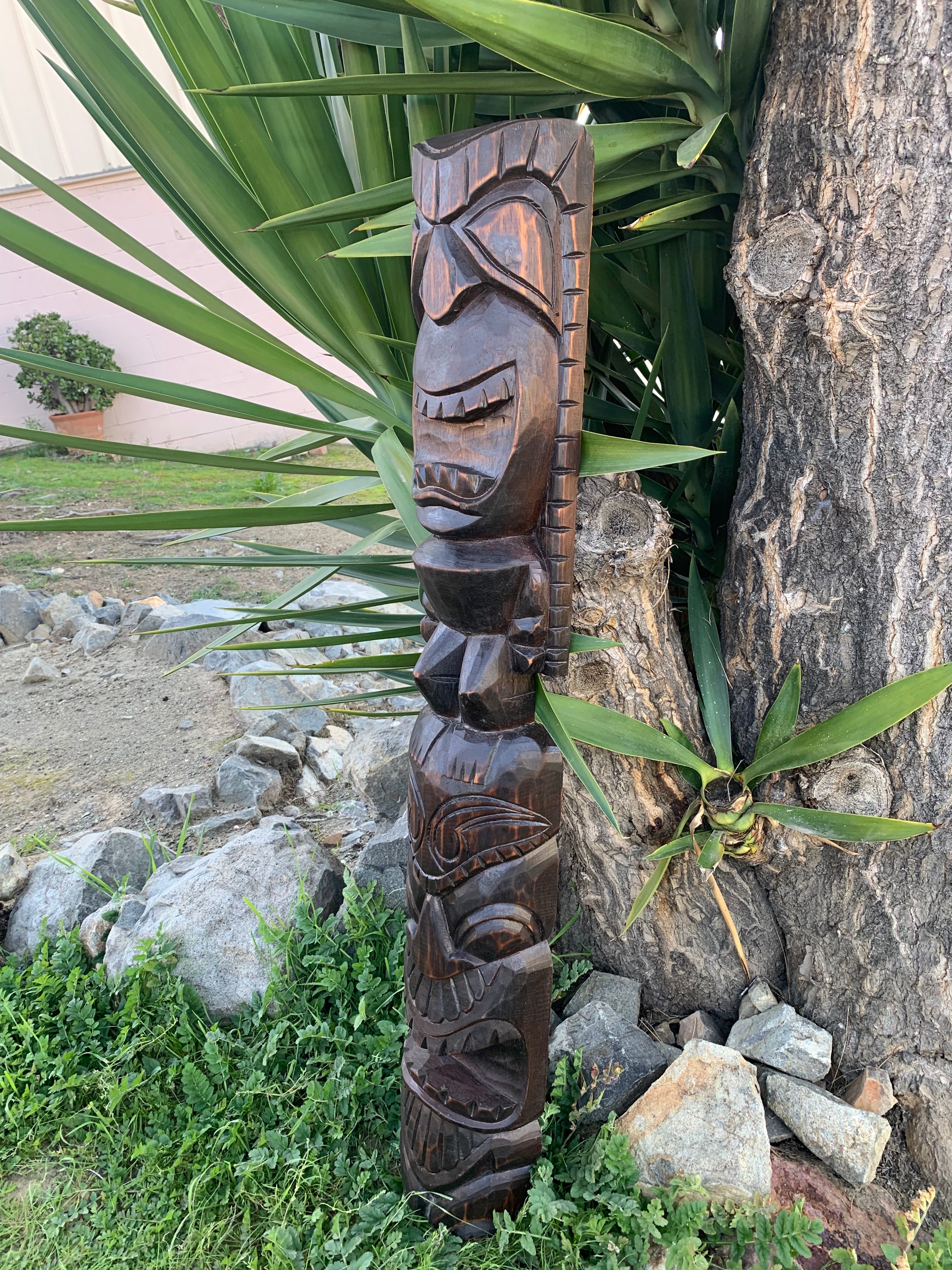 Dark carved wooden tiki totem with faces, placed outdoors beside a tree amid green plants