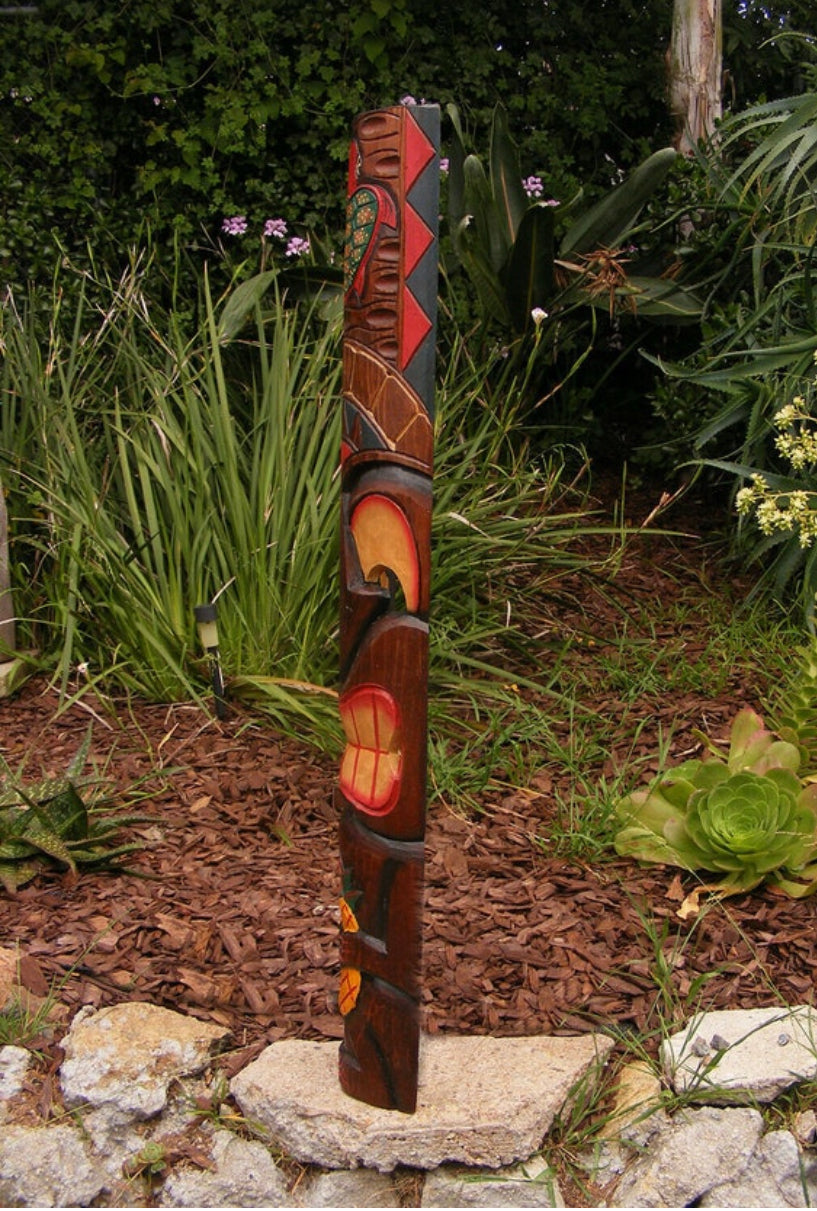 Carved wooden totem pole standing in a garden among mulch, rocks and greenery.