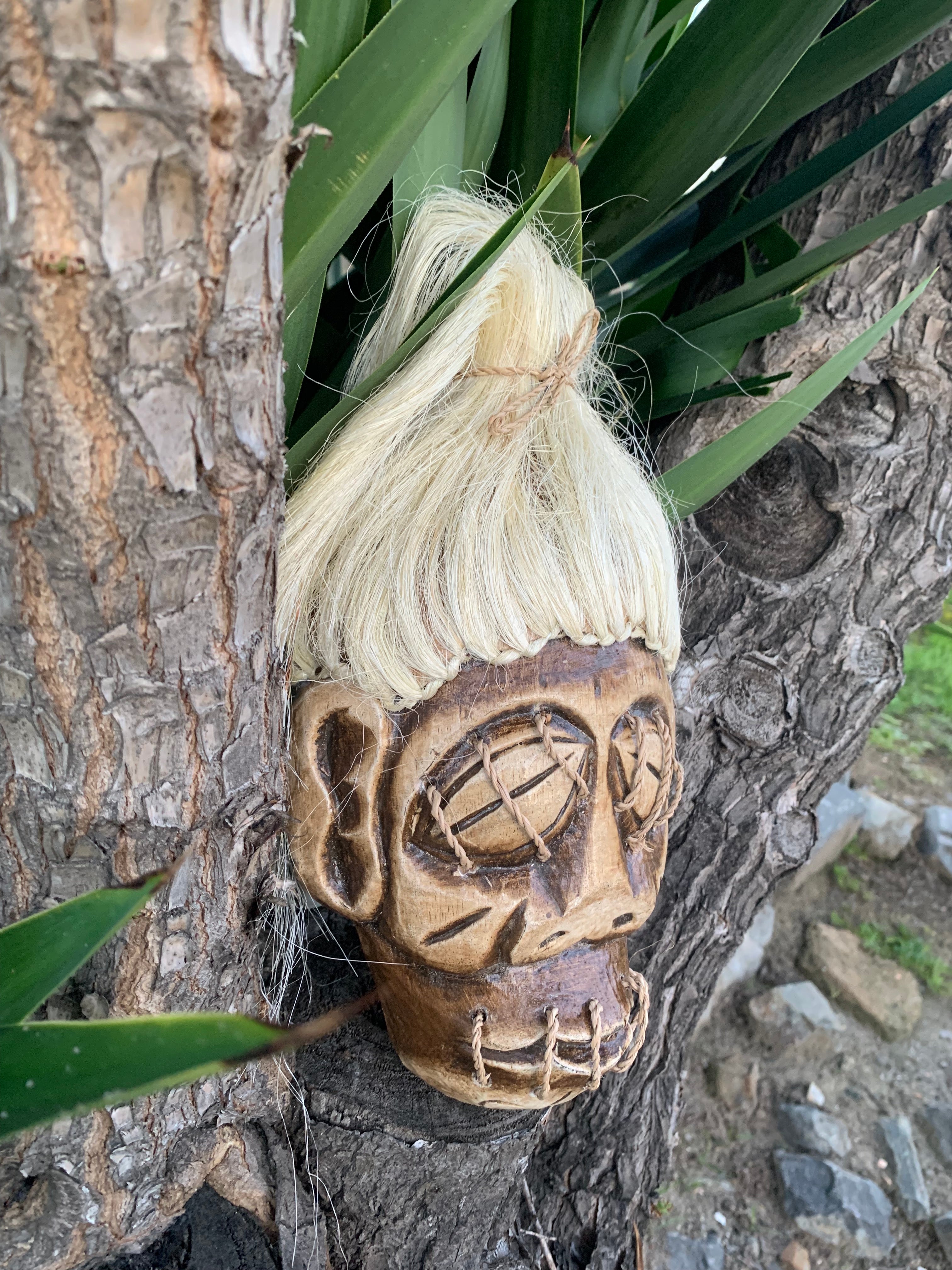 Carved wooden mask with stitched eyes and a blonde wig hanging on a tree trunk