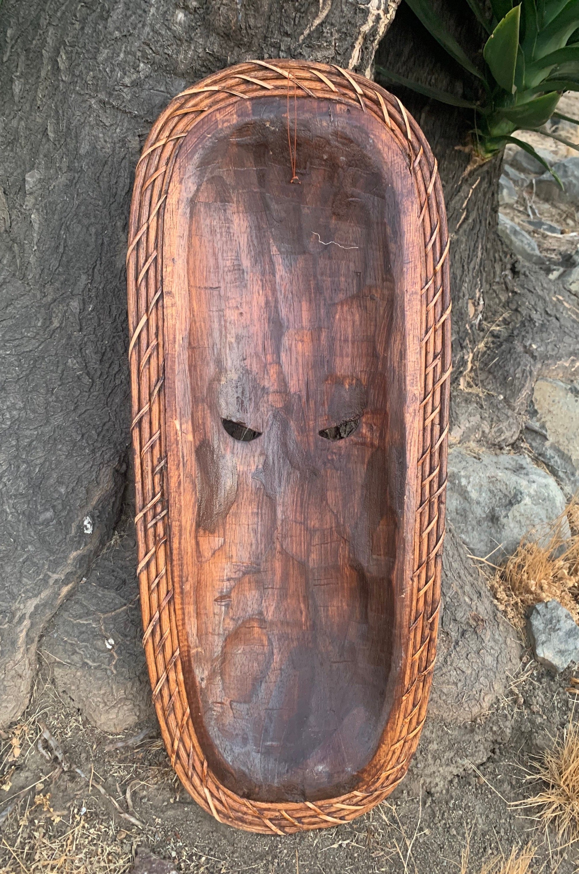 Wooden mask with two eye slits and braided rope border leaning against a tree outdoors