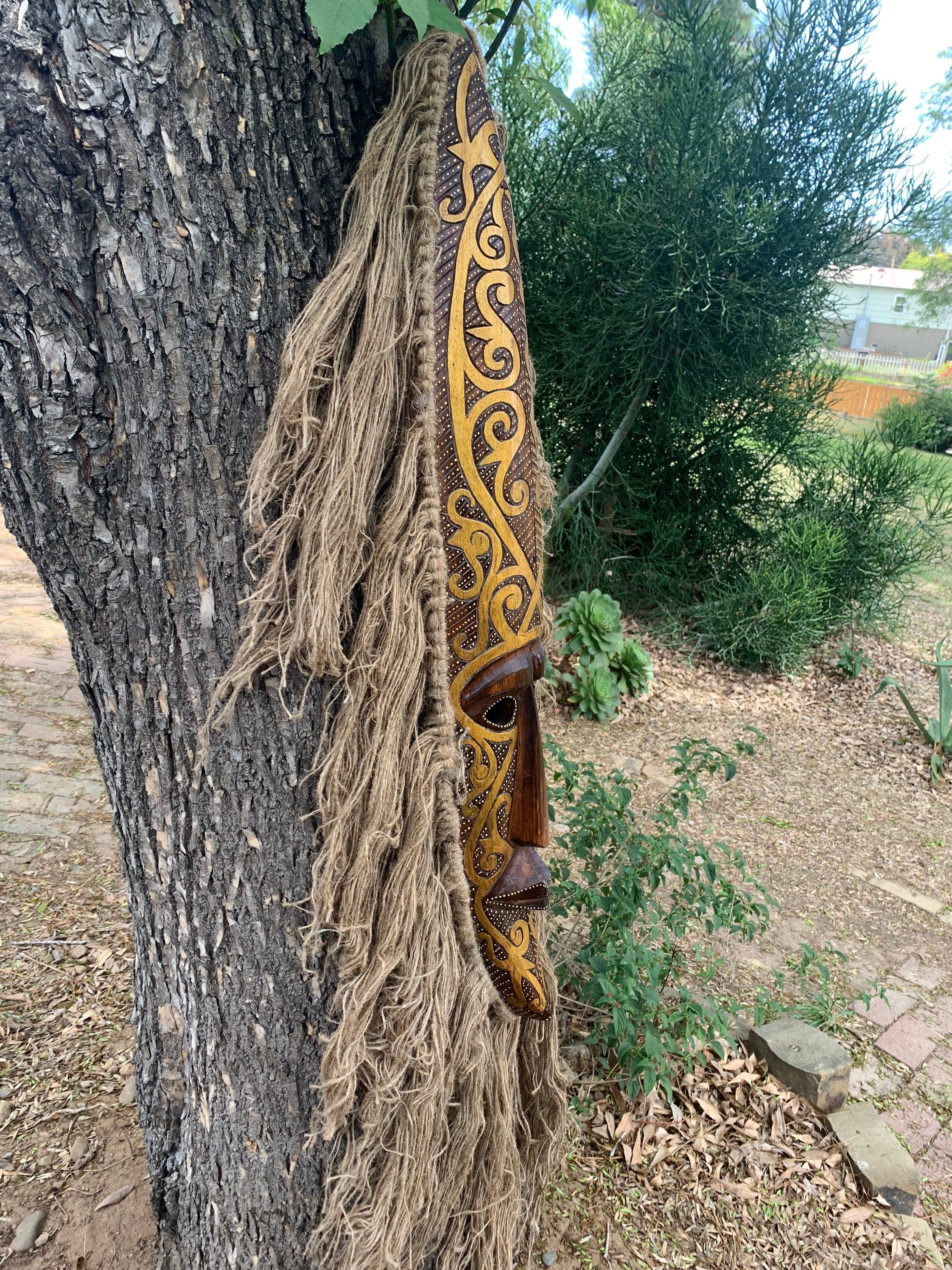 Carved wooden mask with swirling yellow patterns and long beige fiber fringe hanging on a tree.