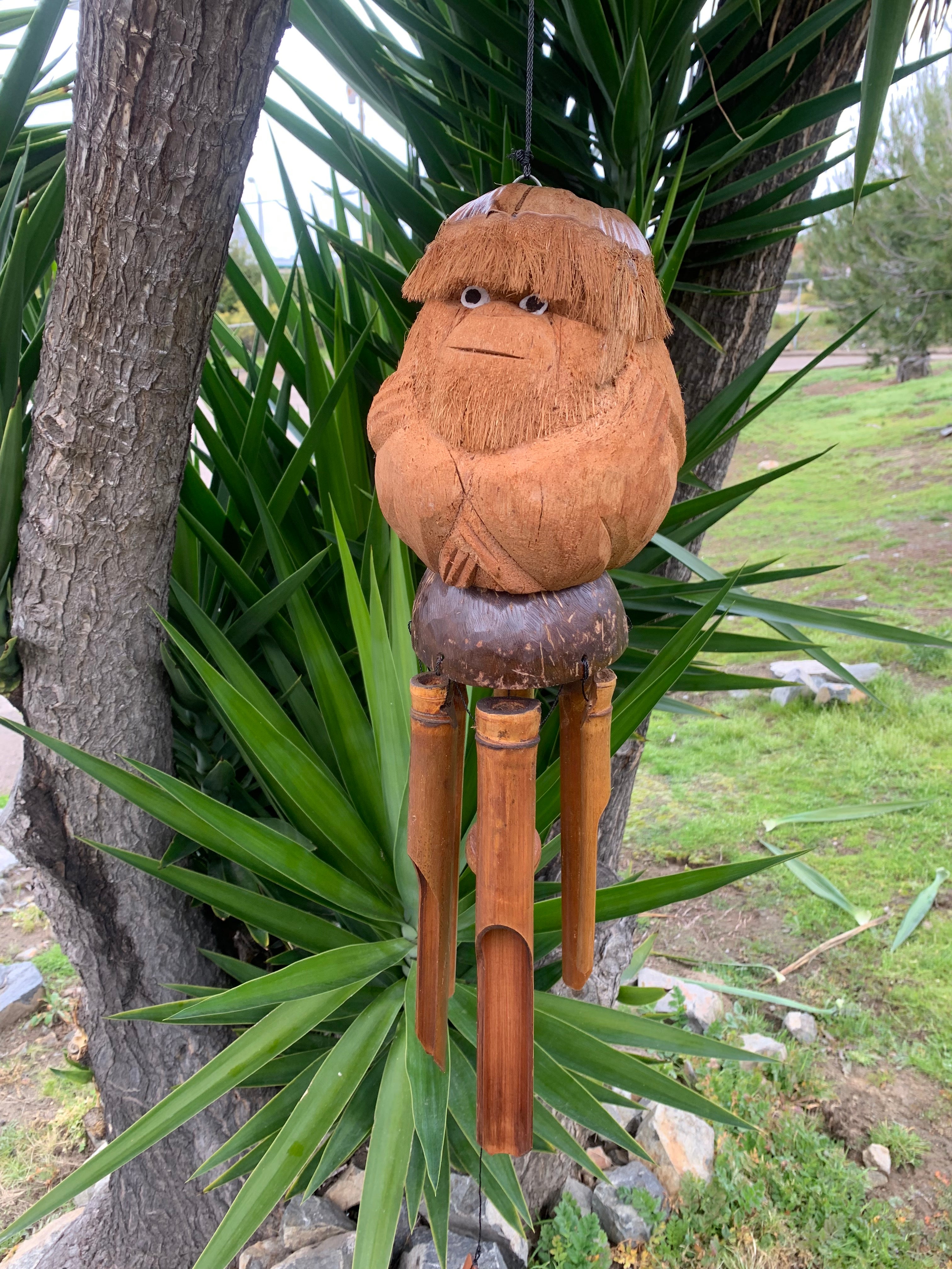 Carved wooden tiki figure with shaggy hair and bamboo legs, hanging in a garden.