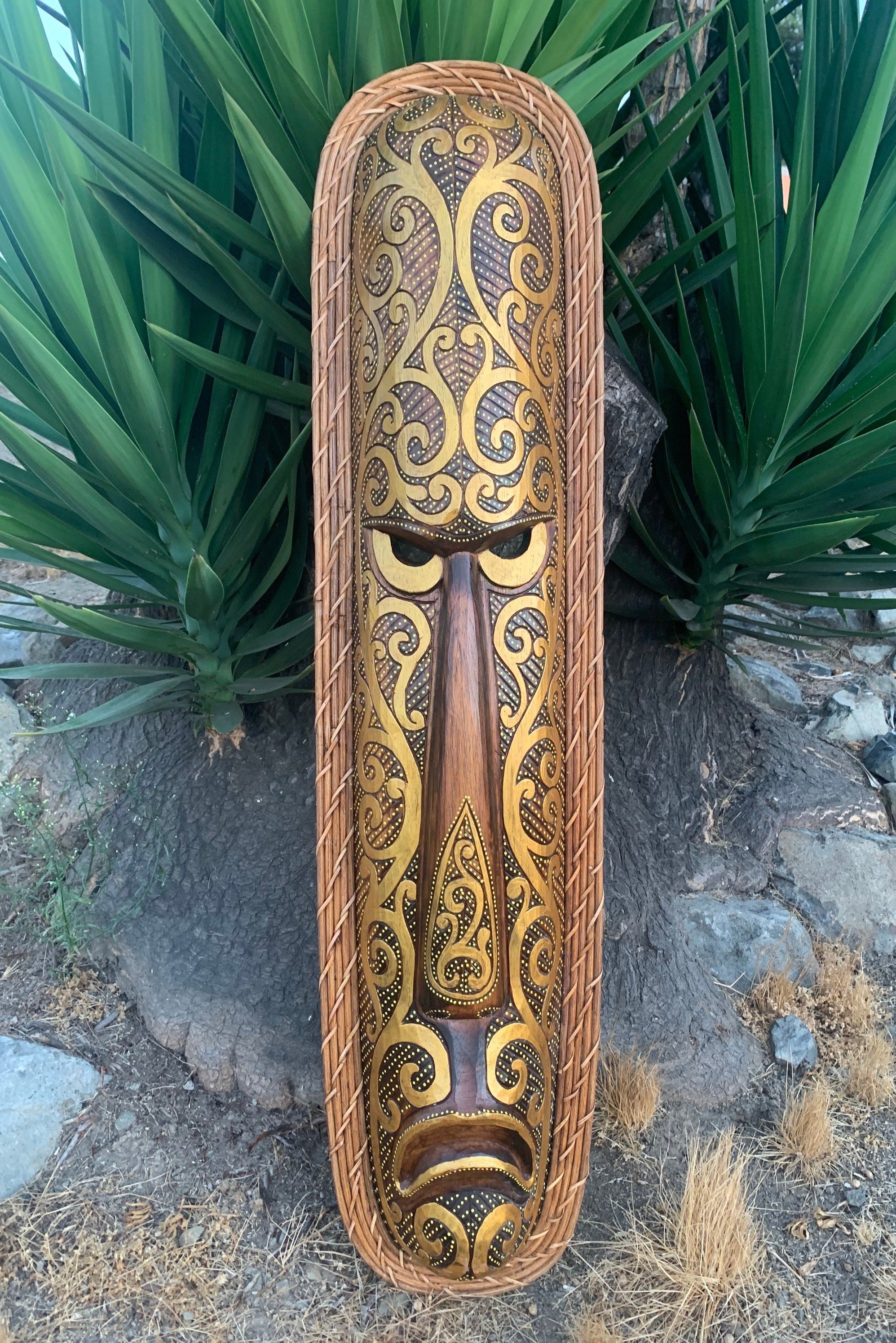 Intricate gold and brown tiki mask with swirling patterns in a woven wicker frame.