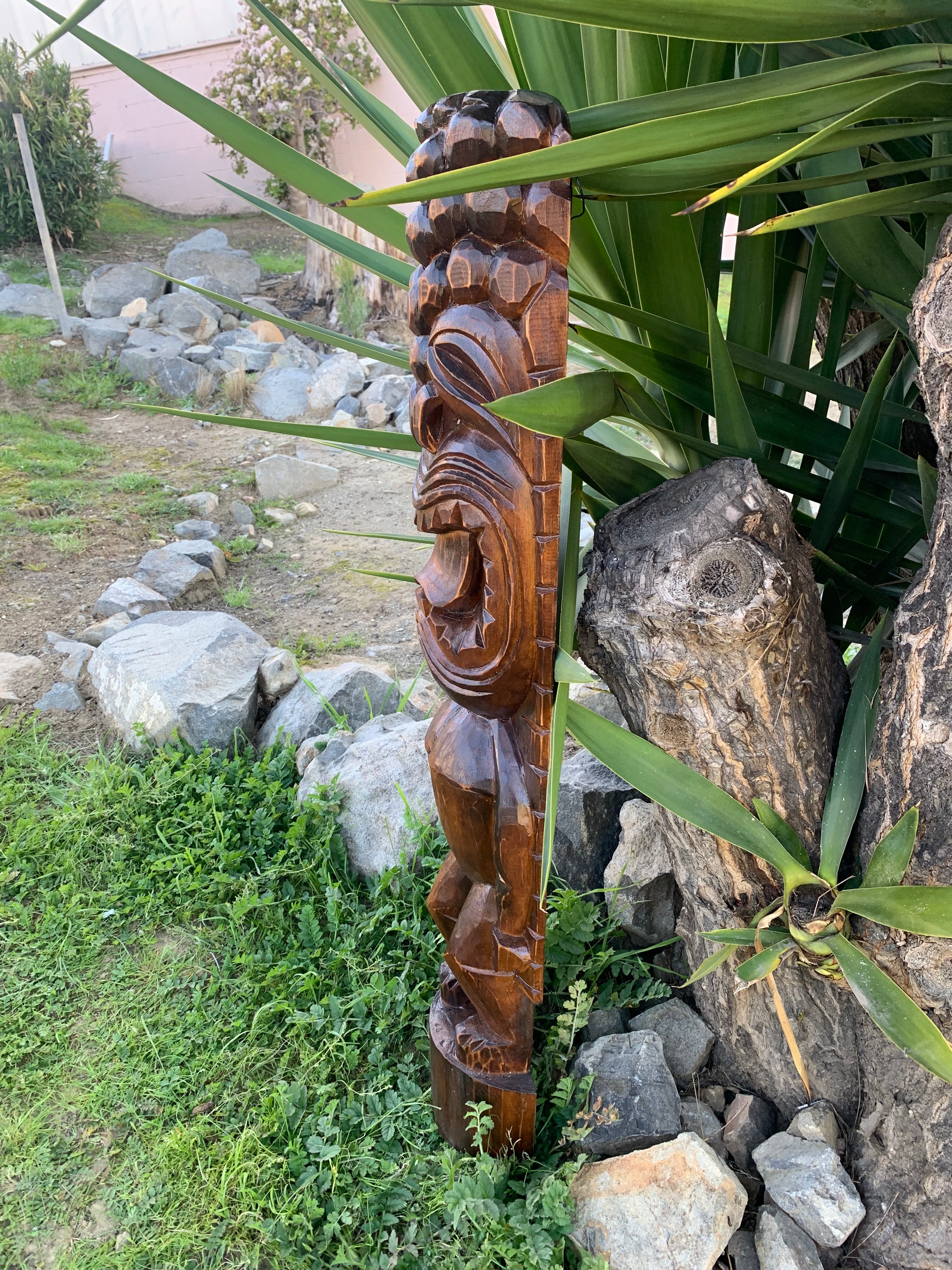 Carved wooden tiki statue standing in a garden with rocks and large green leaves