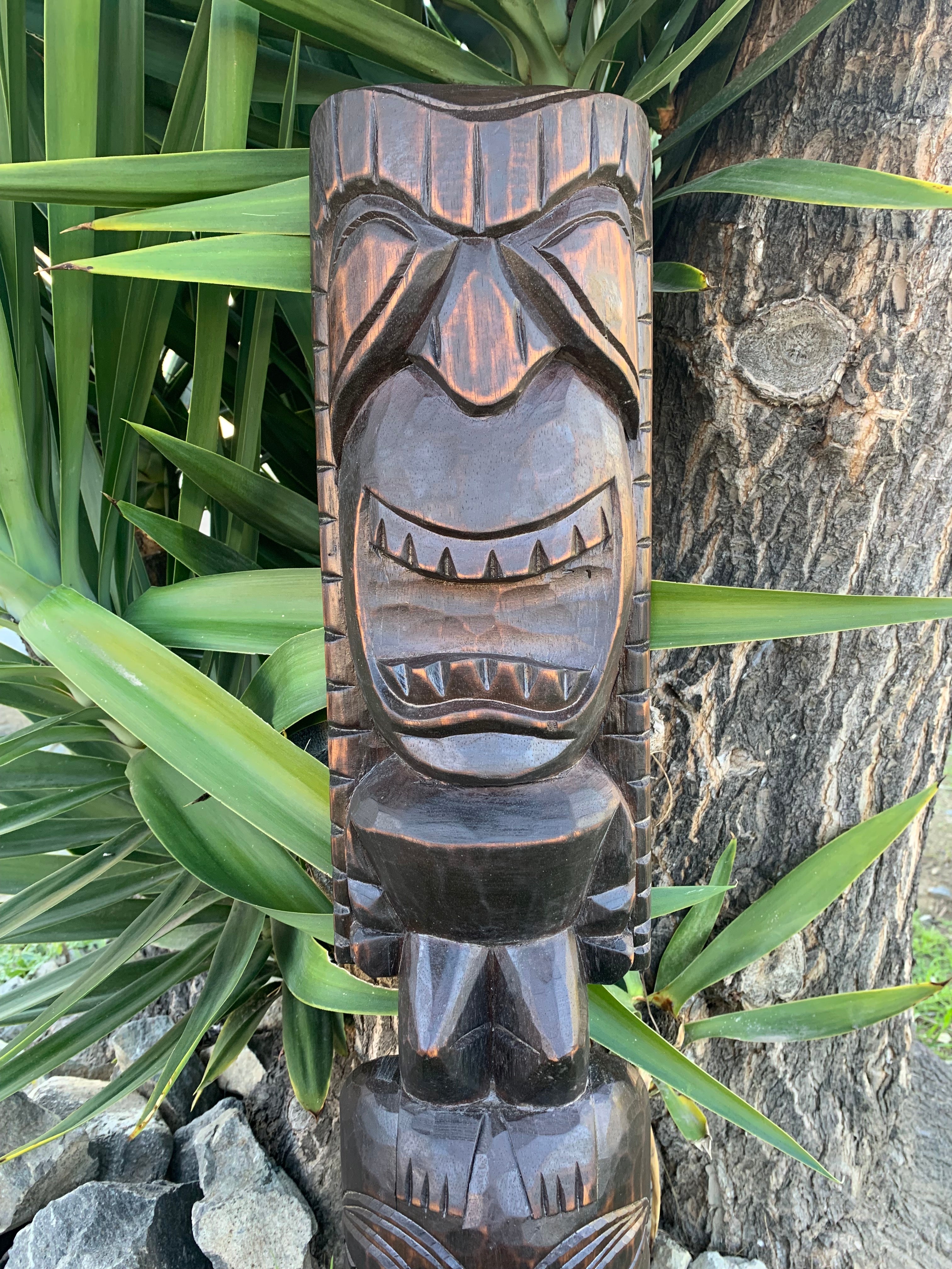 carved wooden tiki statue with a wide grin among green tropical leaves and a tree trunk
