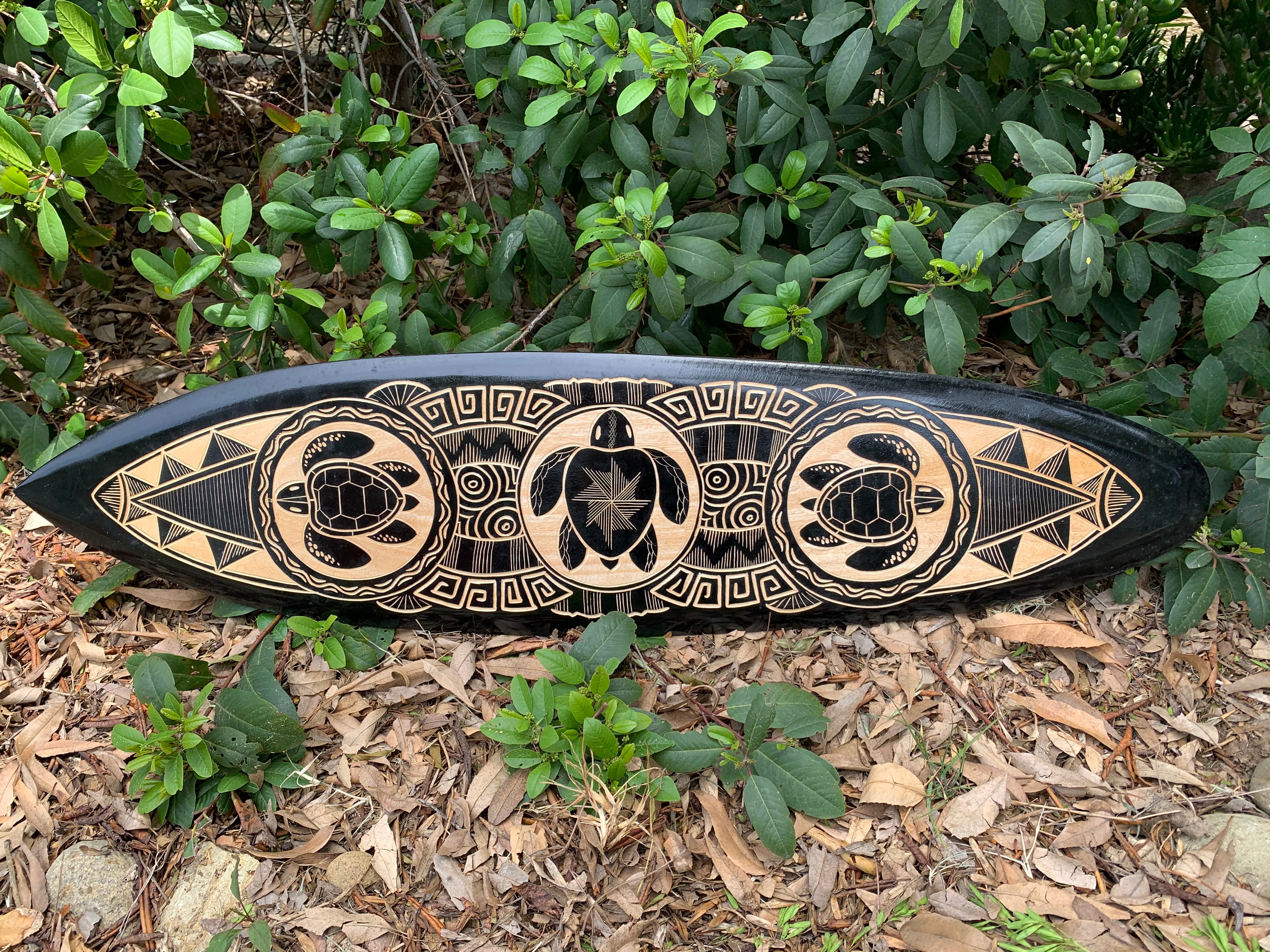 Carved wooden tiki surfboard art on a black surface featuring turtle motifs and geometric patterns
