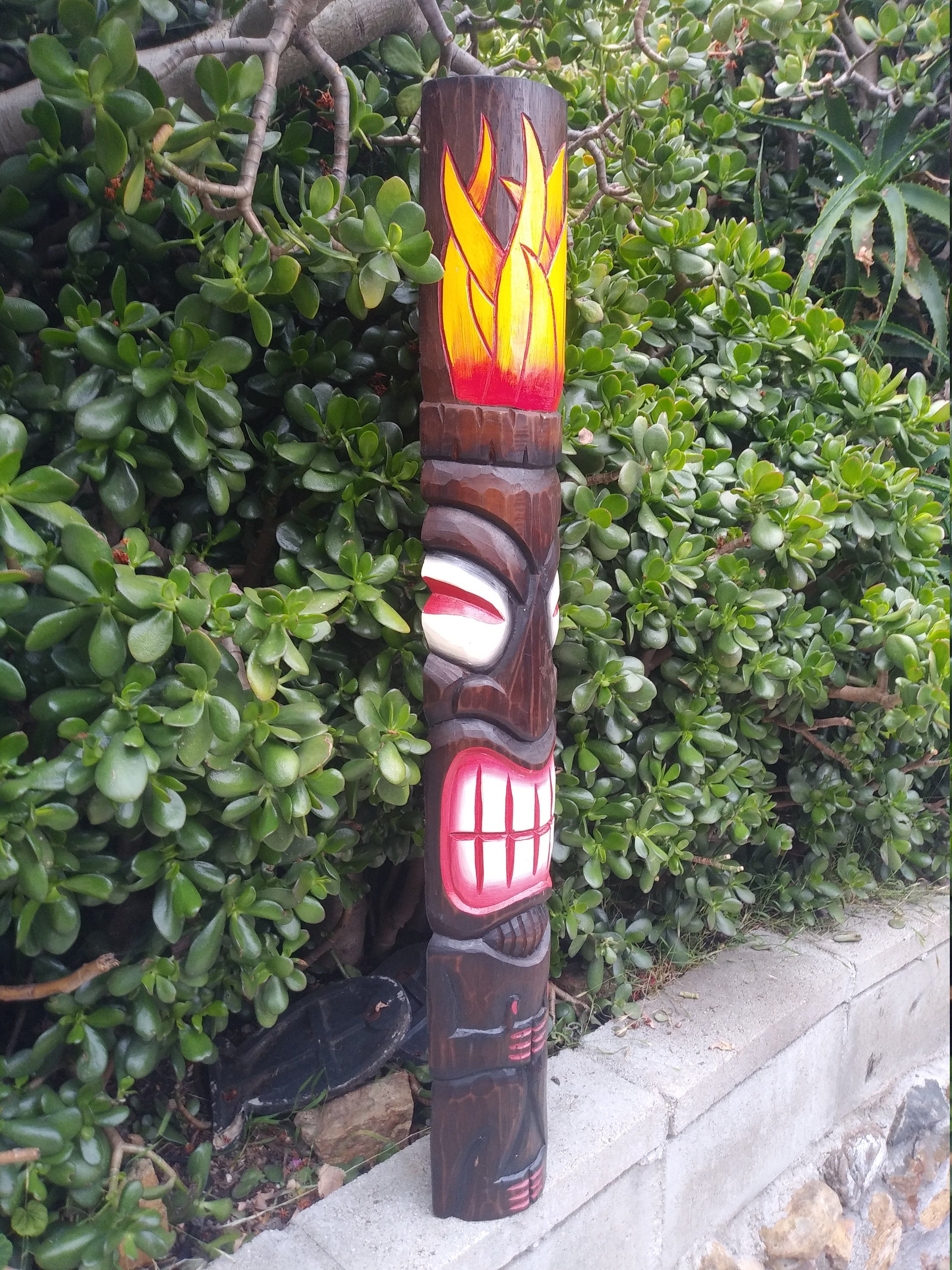 Carved wooden tiki totem pole with flame-like artwork at the top in a garden setting