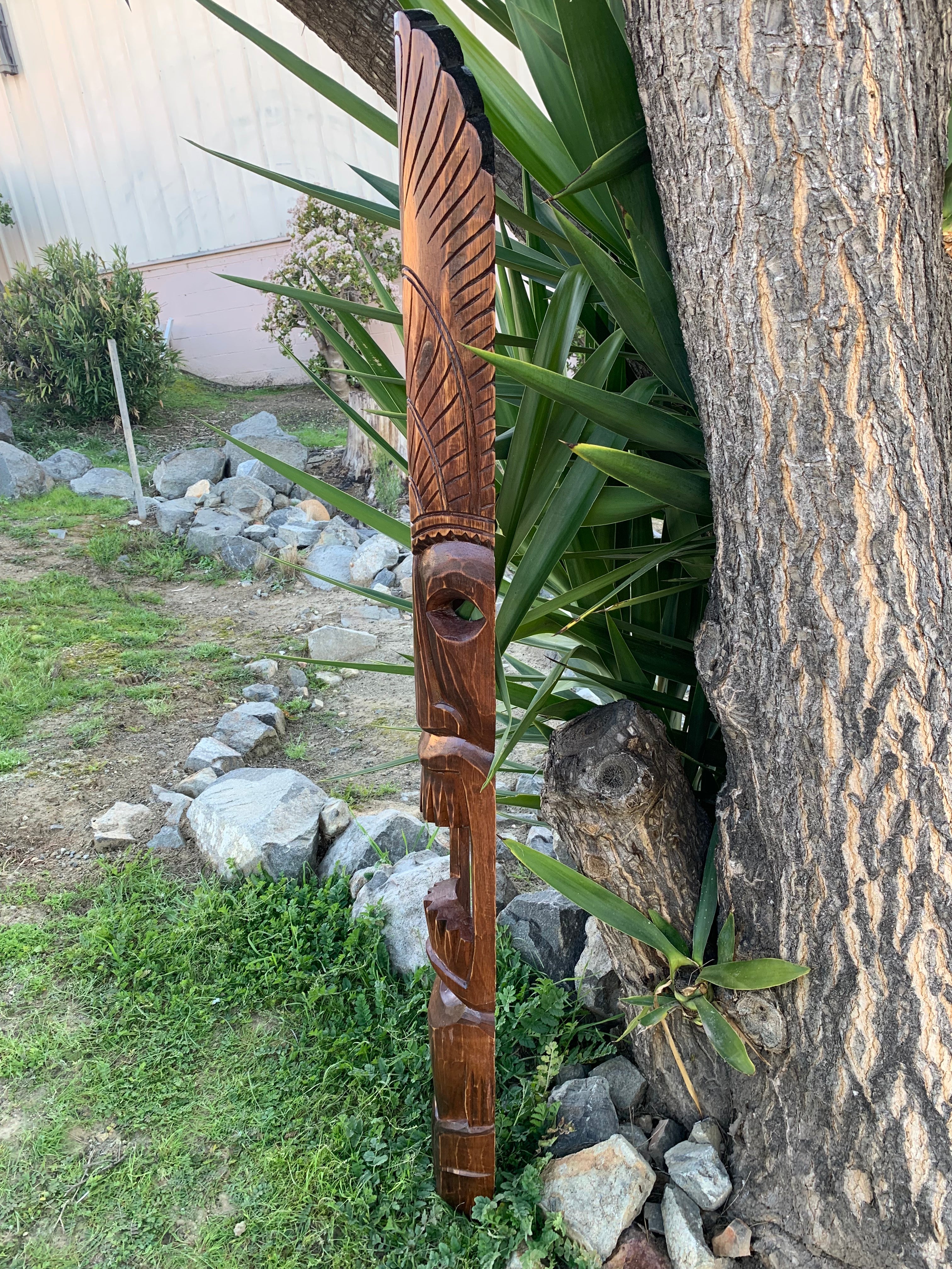 Carved wooden totem pole with feather-like patterns, leaning against a tree in a garden.