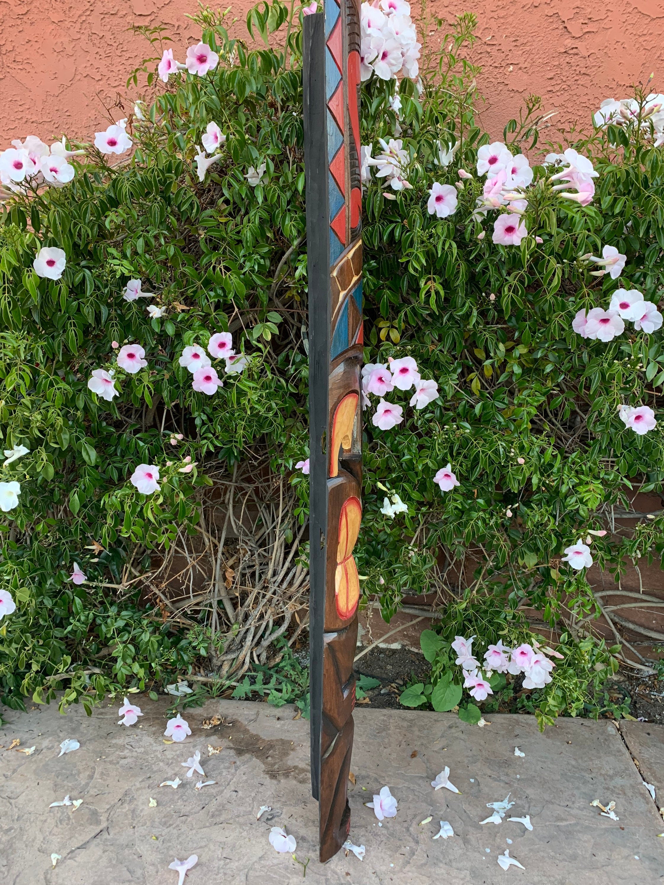 Vertical carved wooden totem pole beside pink and white hibiscus flowers in a garden.