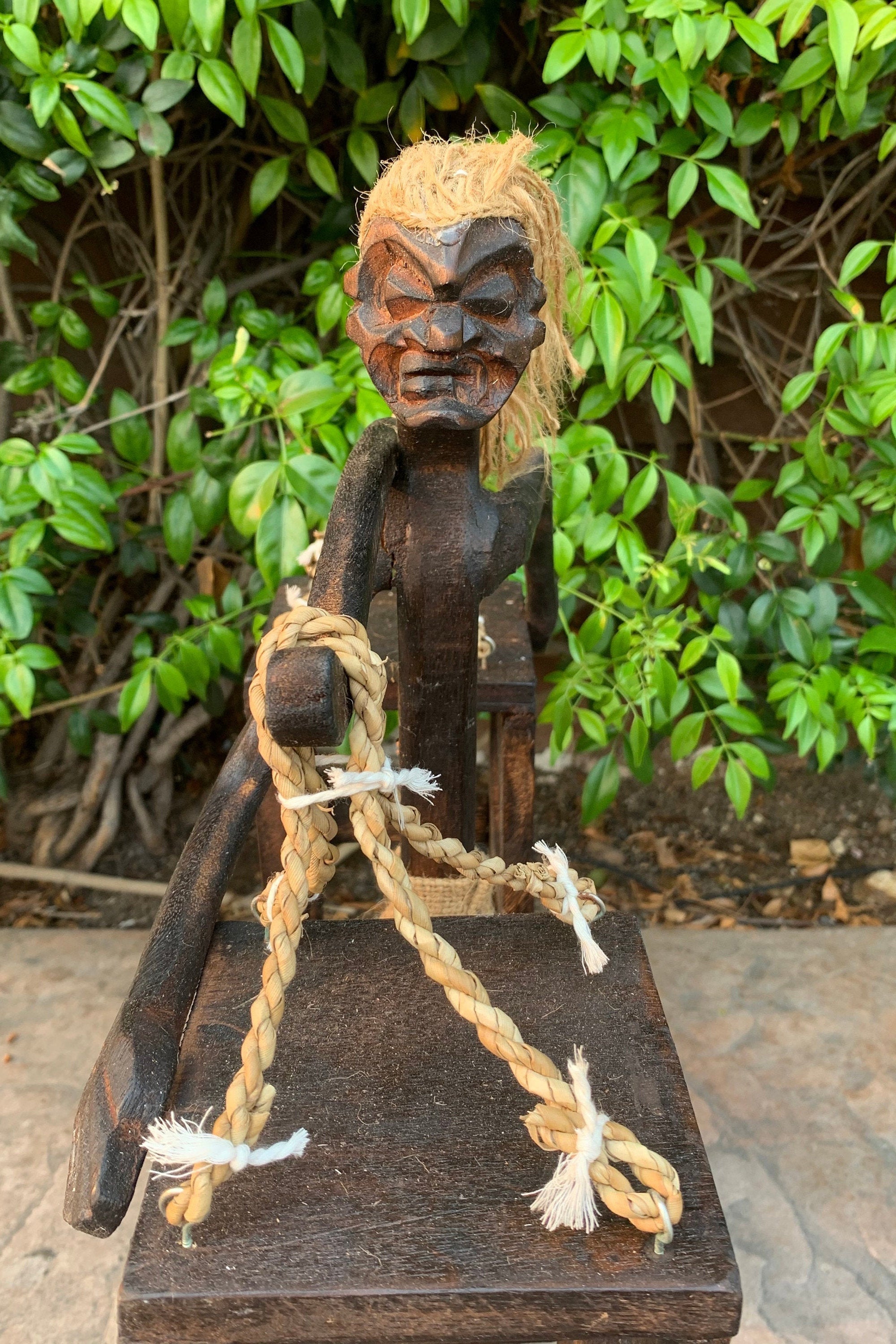 Carved wooden tribal statue with straw hair and coiled rope on a pedestal in a garden.