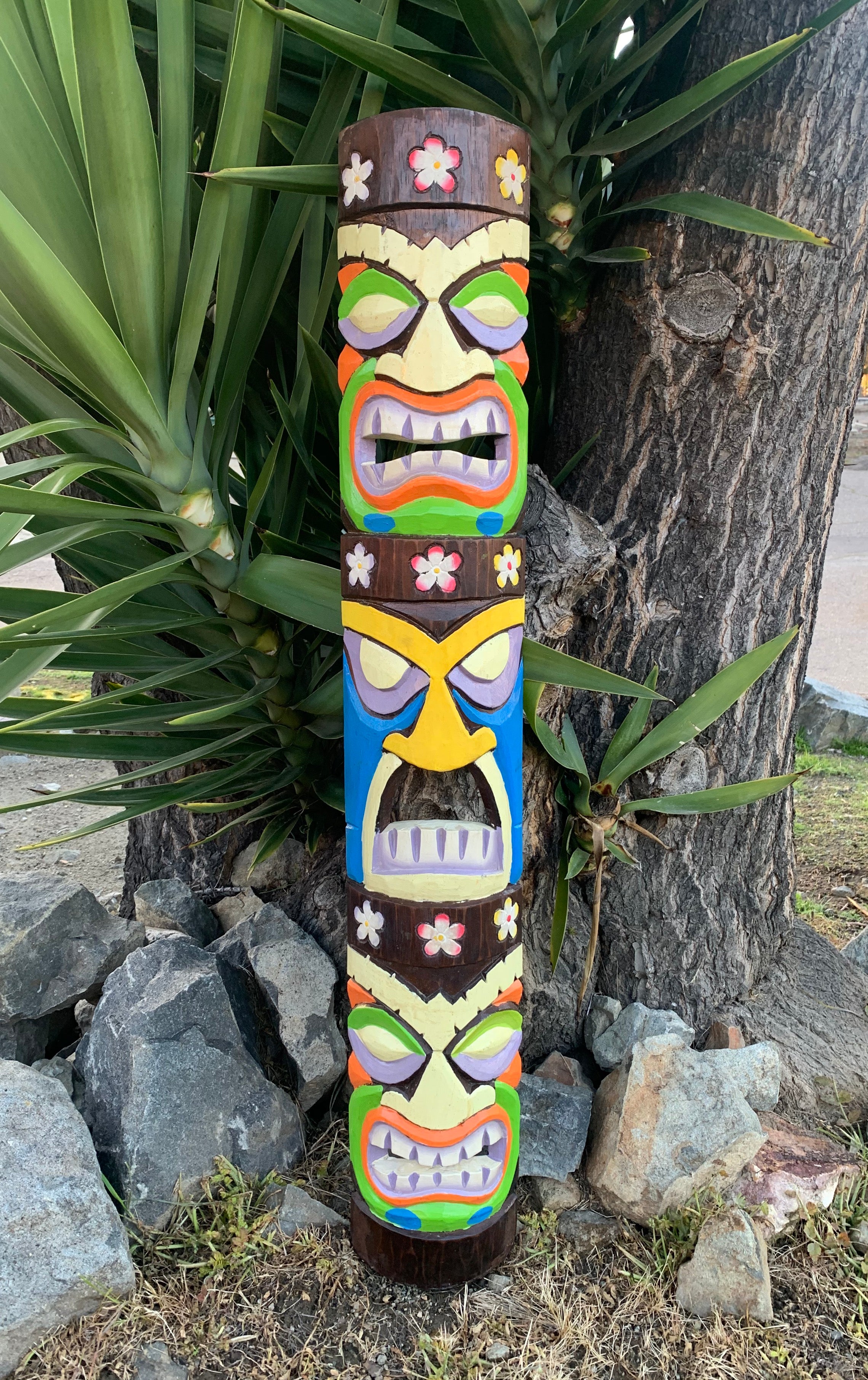 Colorful carved tiki totem pole with multiple faces and flower motifs beside a tree.