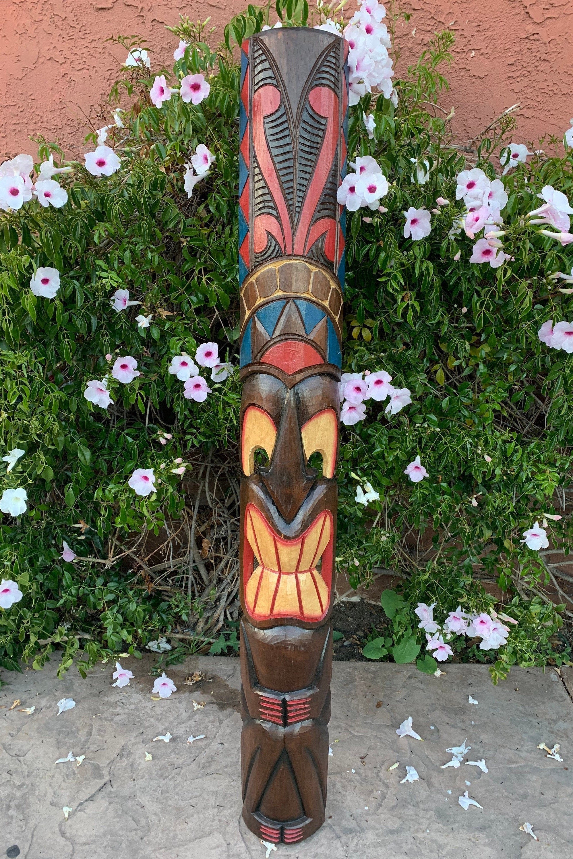 Colorful carved wooden totem pole with geometric patterns in a garden of pink and white flowers