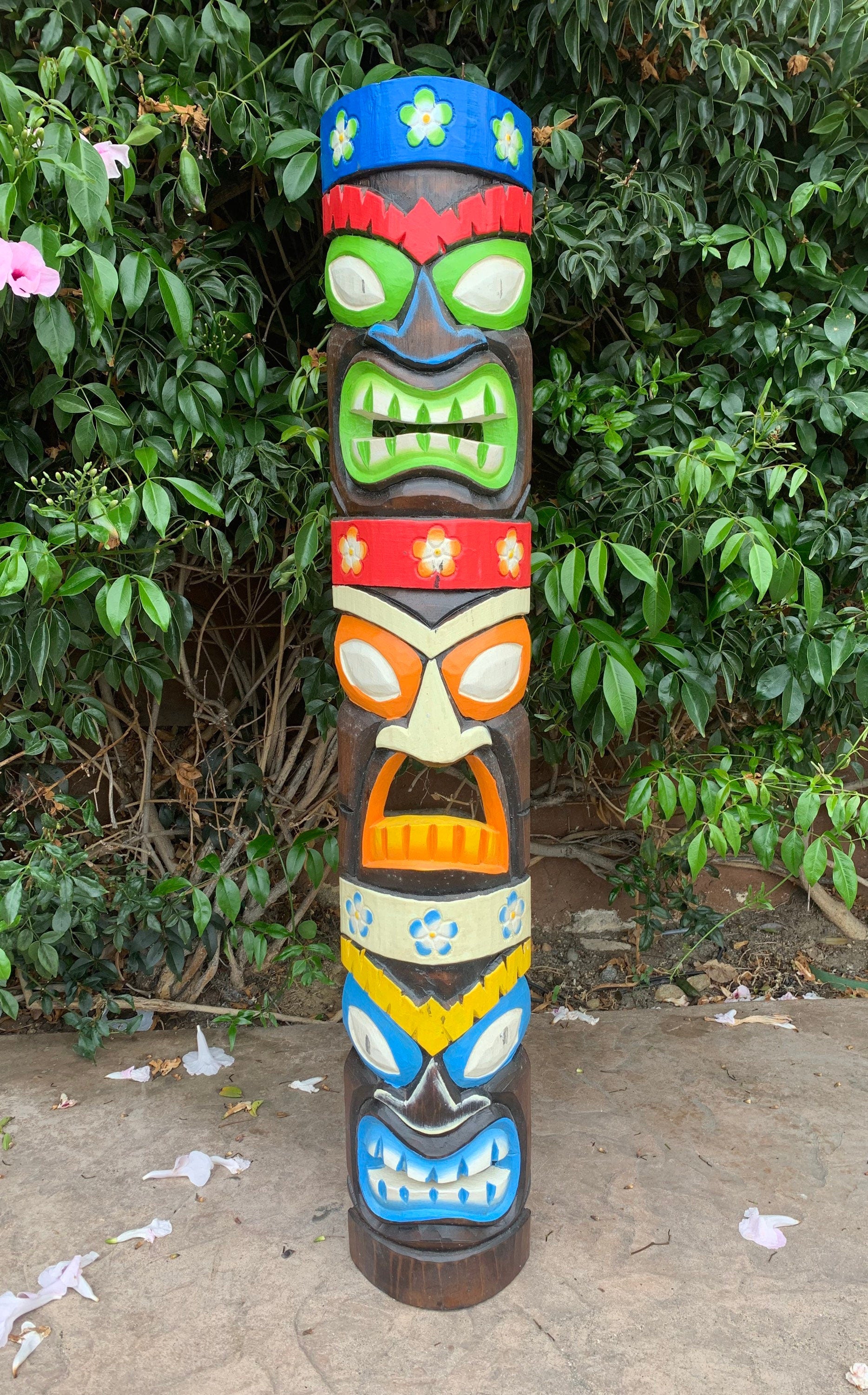 Colorful carved totem pole with stacked tiki faces outdoors against leafy bushes
