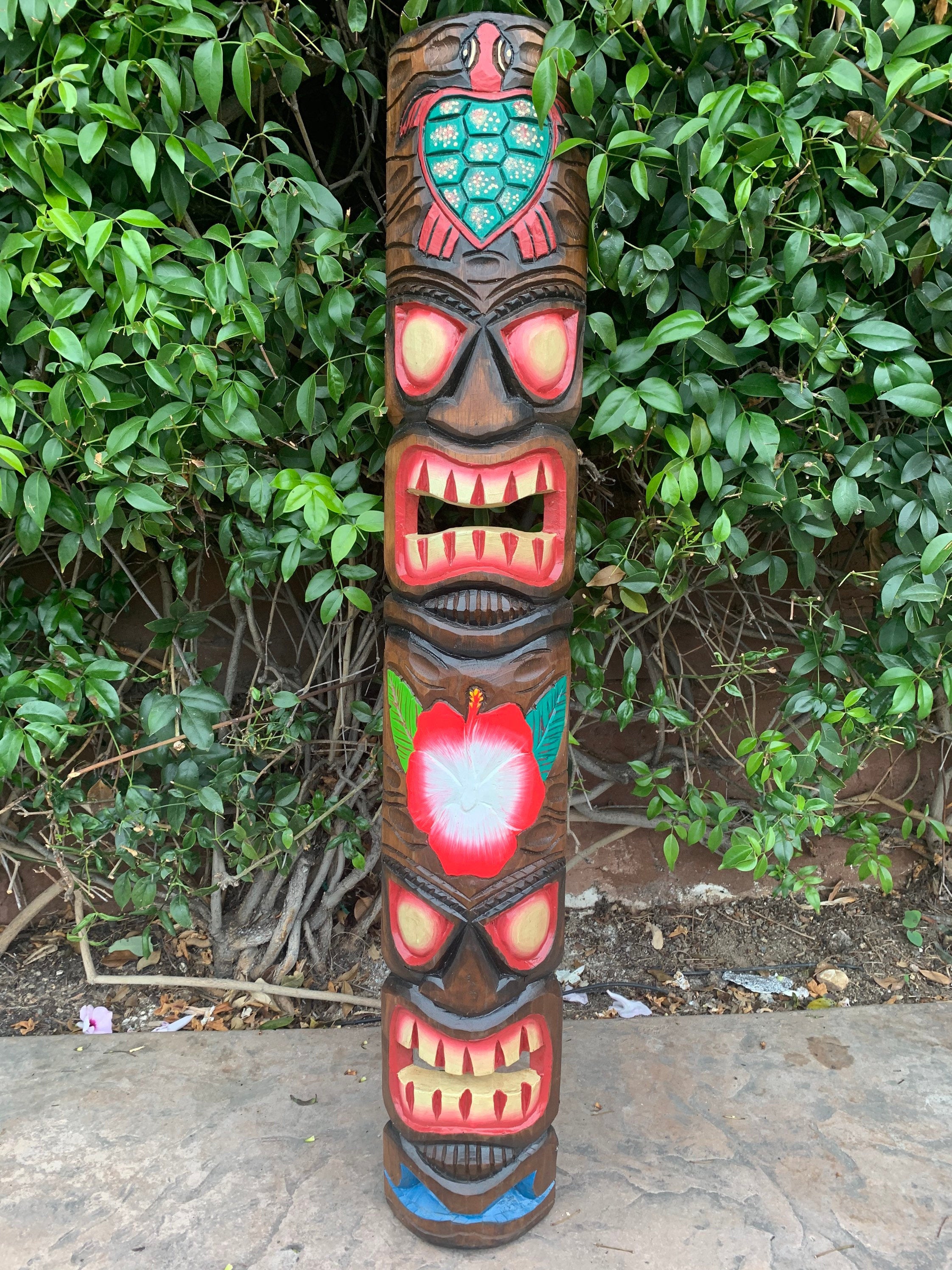 Colorful carved totem pole with multiple faces and a hibiscus flower, set against green foliage.