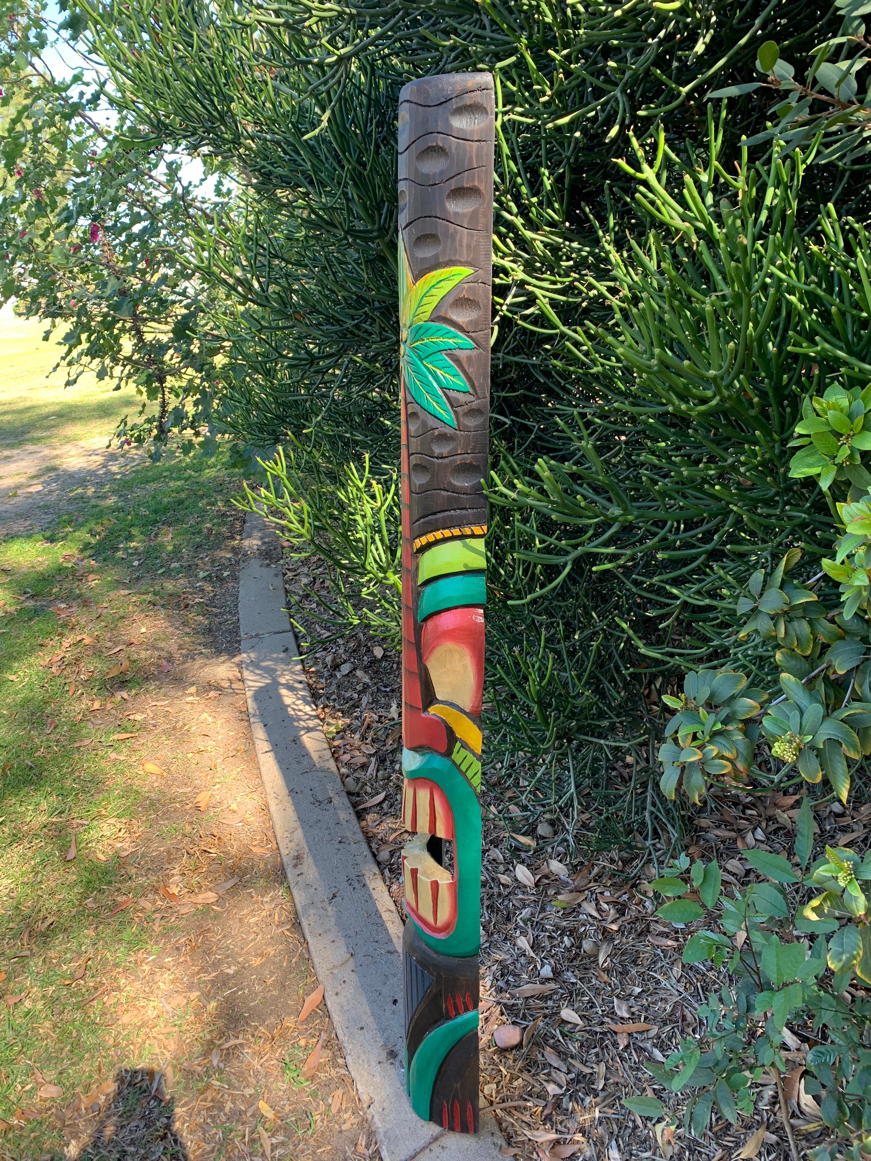 Colorful carved wooden totem pole with leaf motif beside a hedge in a sunny garden