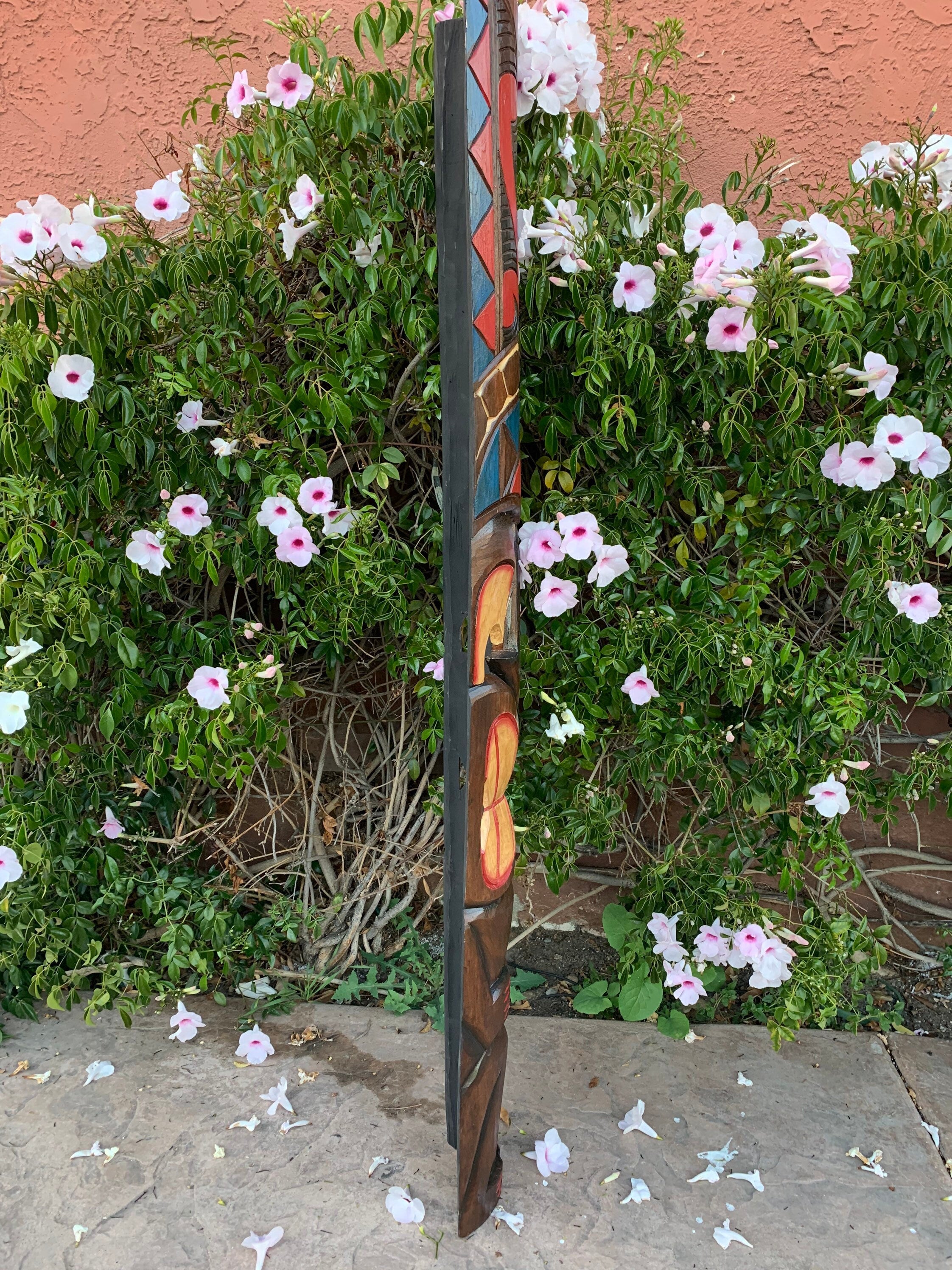 Colorful carved totem pole beside a flowering bush with white petals and pink centers.