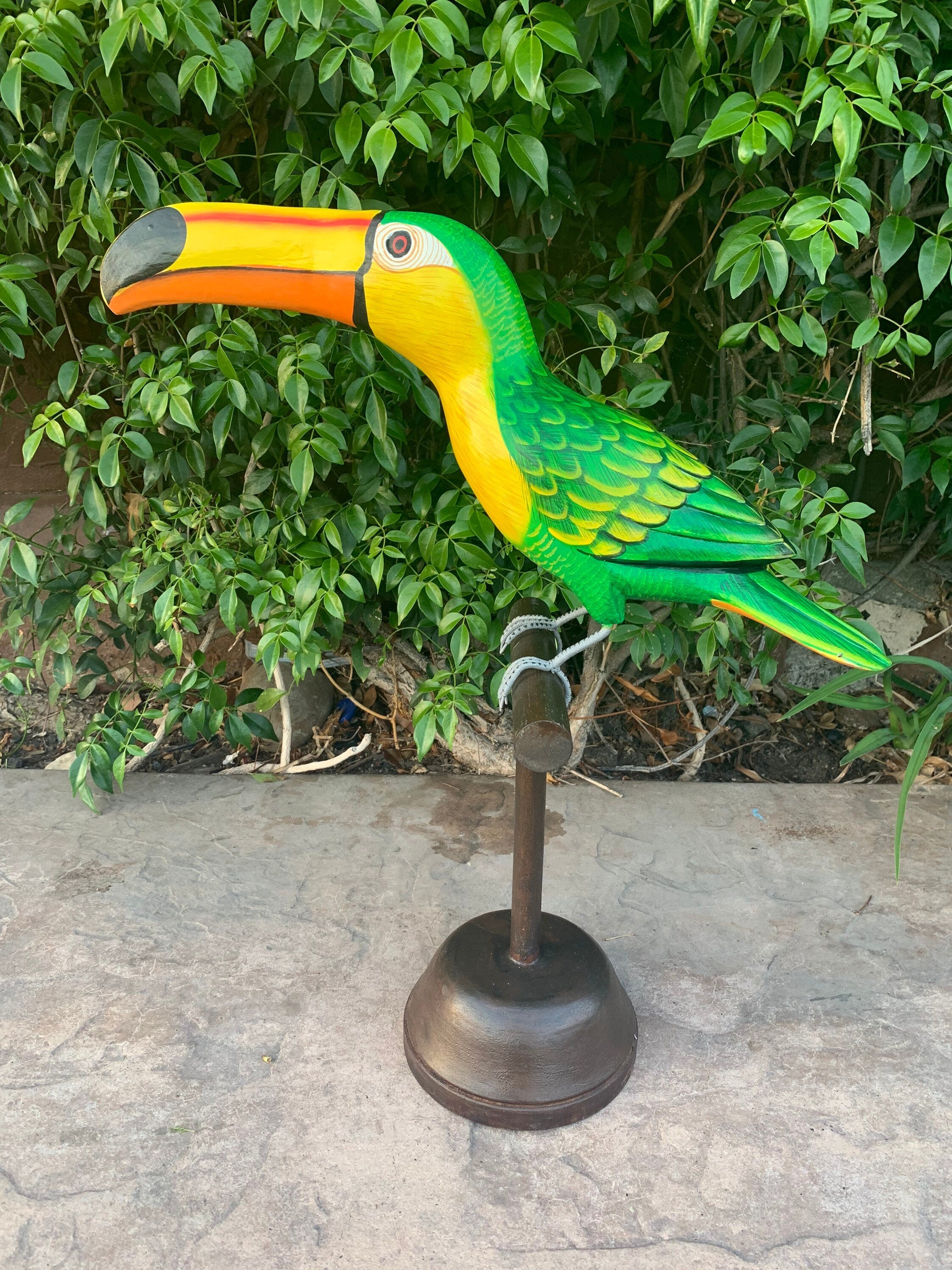 Colorful macaw sculpture with green wings and yellow chest on a metal stand, leafy background