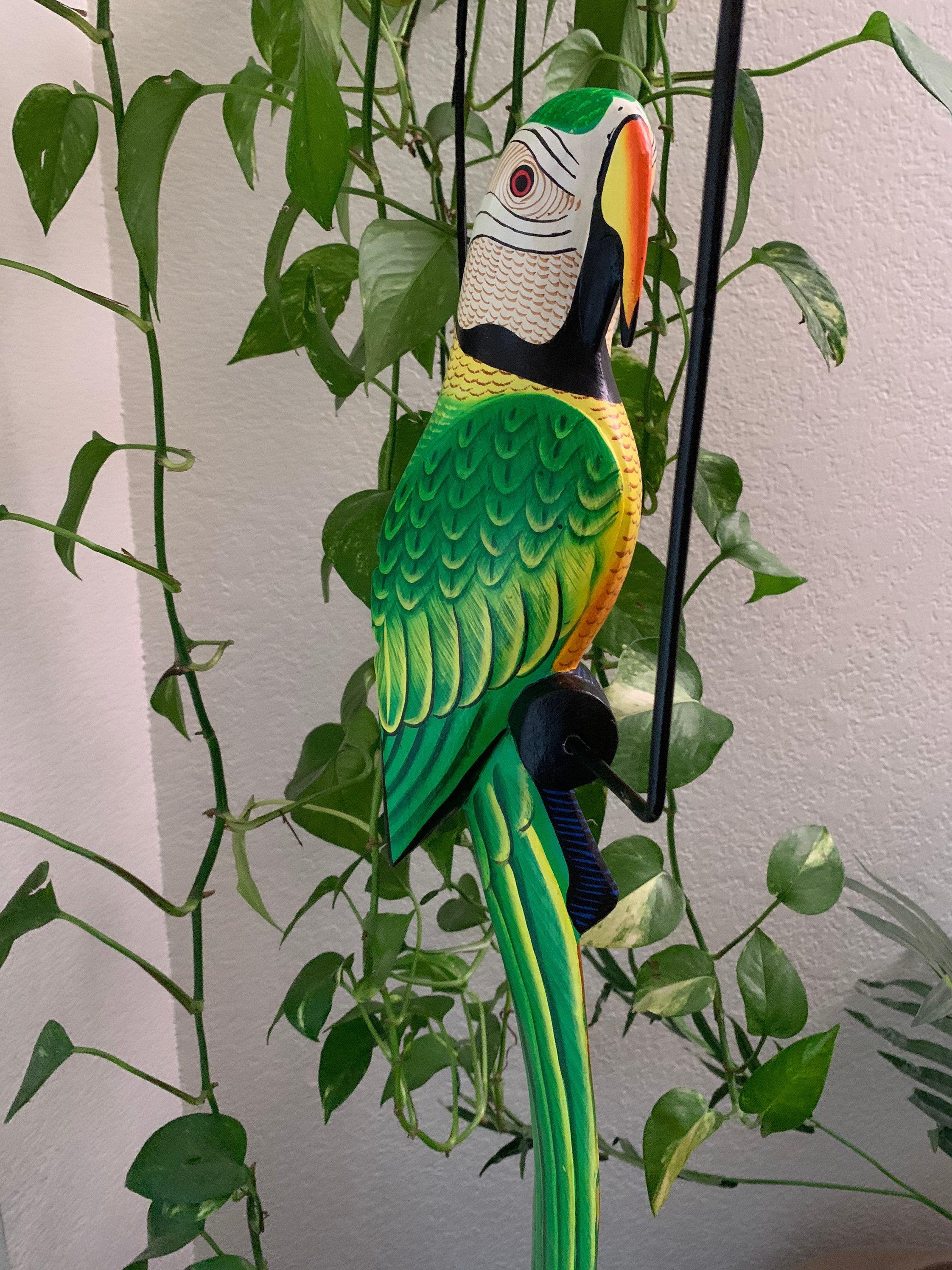 Bright green, yellow, and orange macaw wooden sculpture perched on a stand among houseplants.