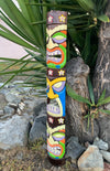 Tall wooden tiki totem pole with multi-colored carved faces and flower accents, displayed outdoors among plants and rocks.