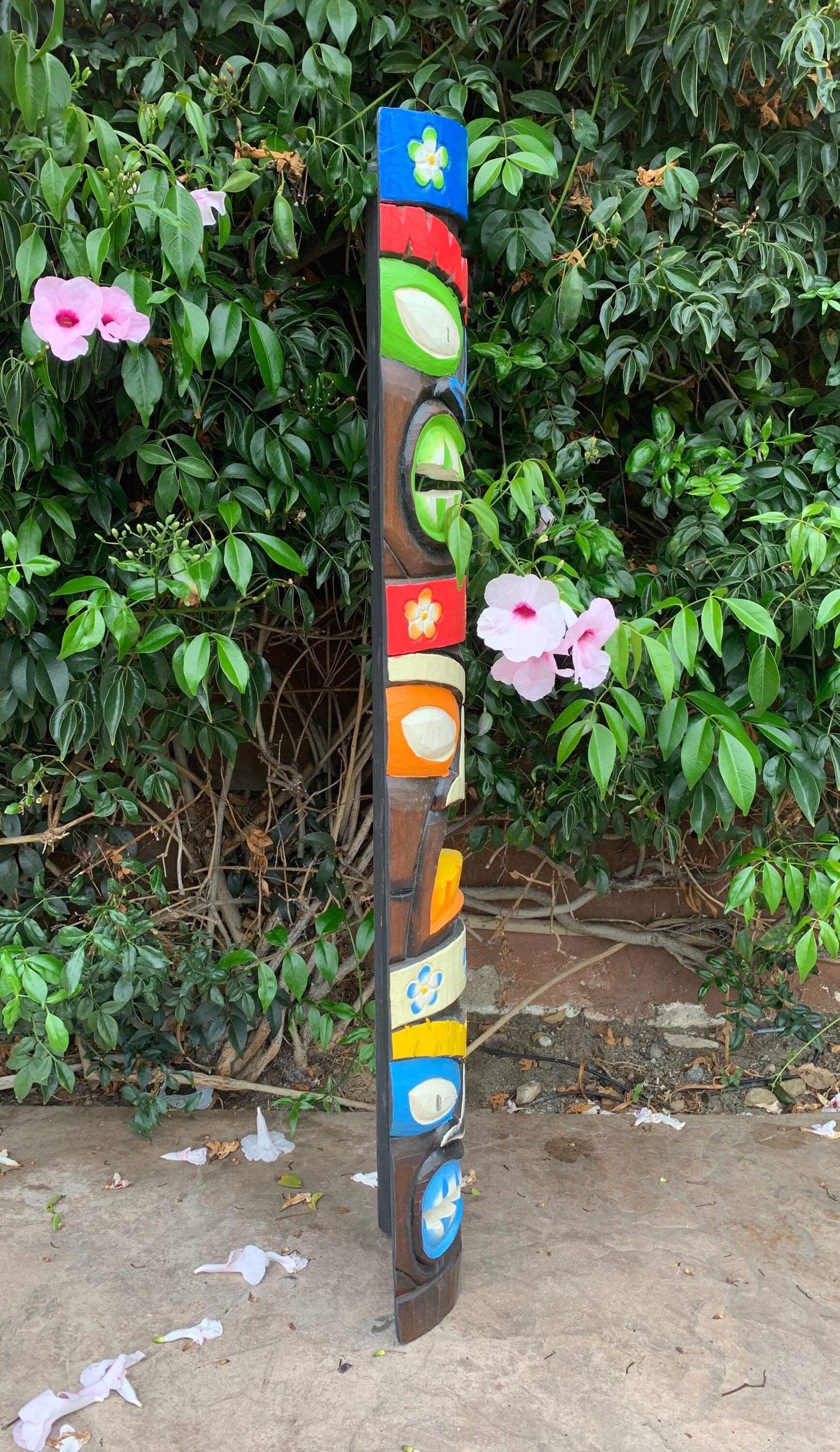 Colorful painted totem pole outdoors beside a green hedge and pink flowers.