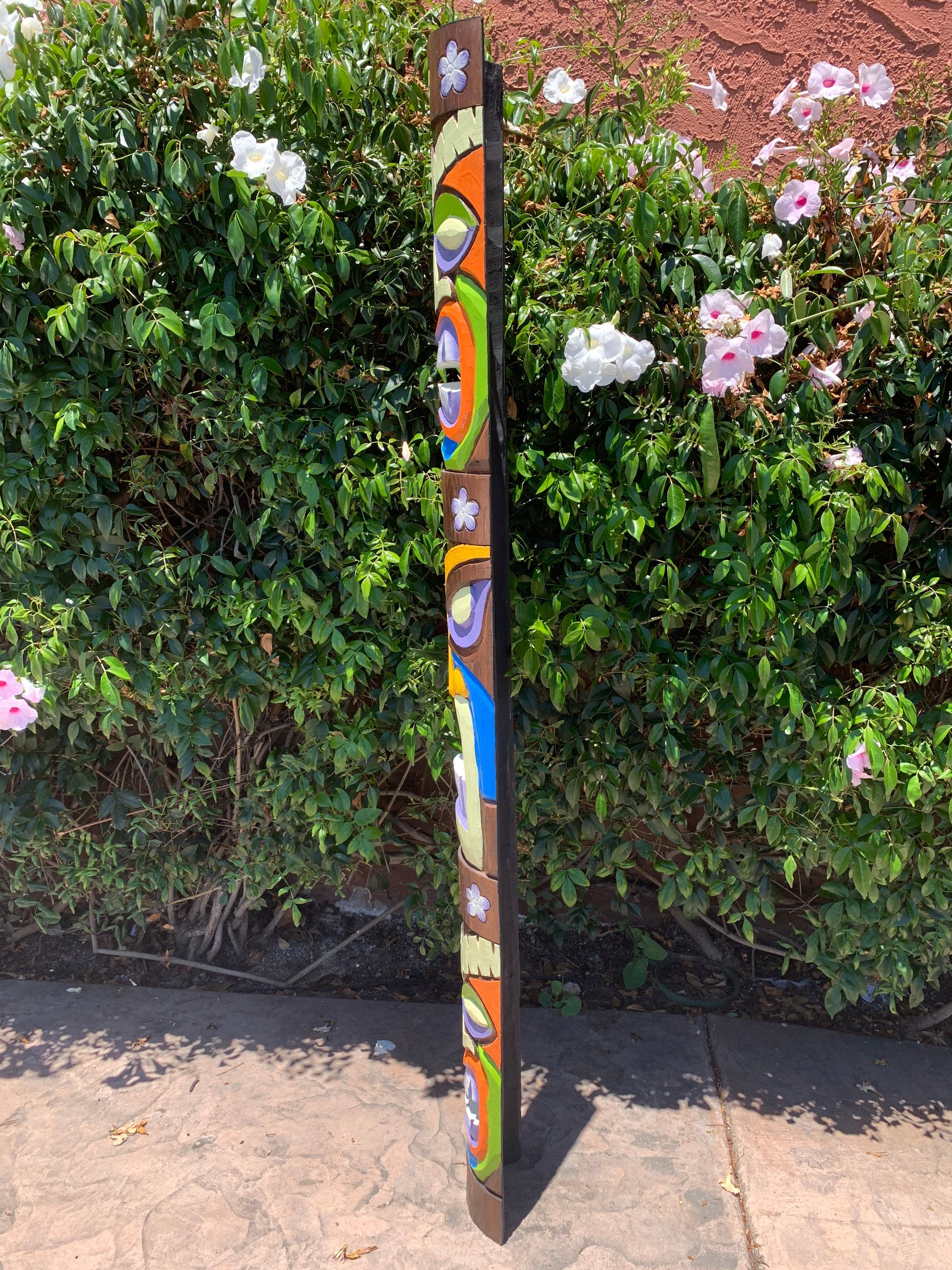 Colorful painted totem pole with geometric patterns beside green shrubs on a sidewalk.