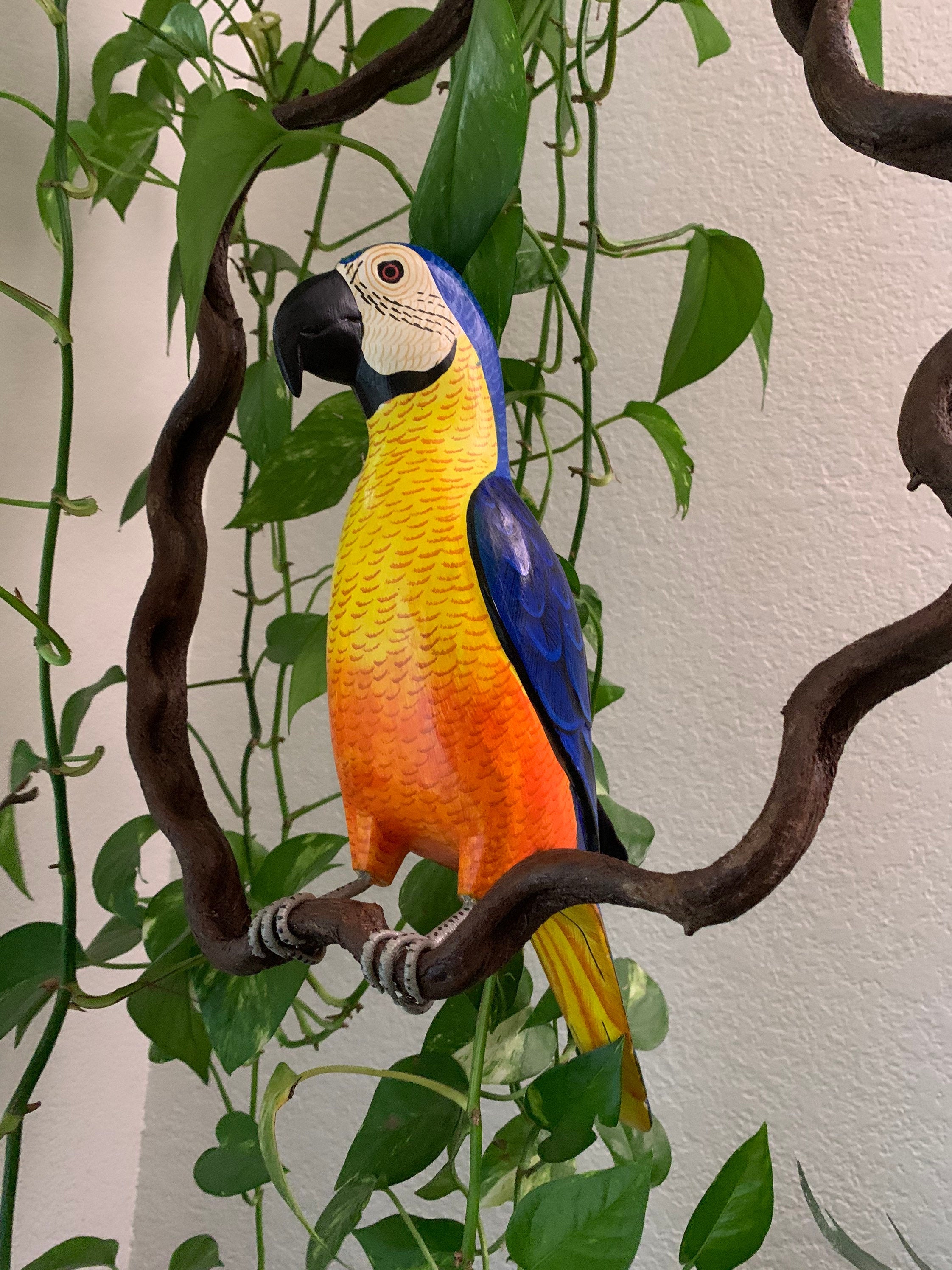 Colorful parrot sculpture with blue wings and yellow-orange chest perched on a twisting branch among green leaves.