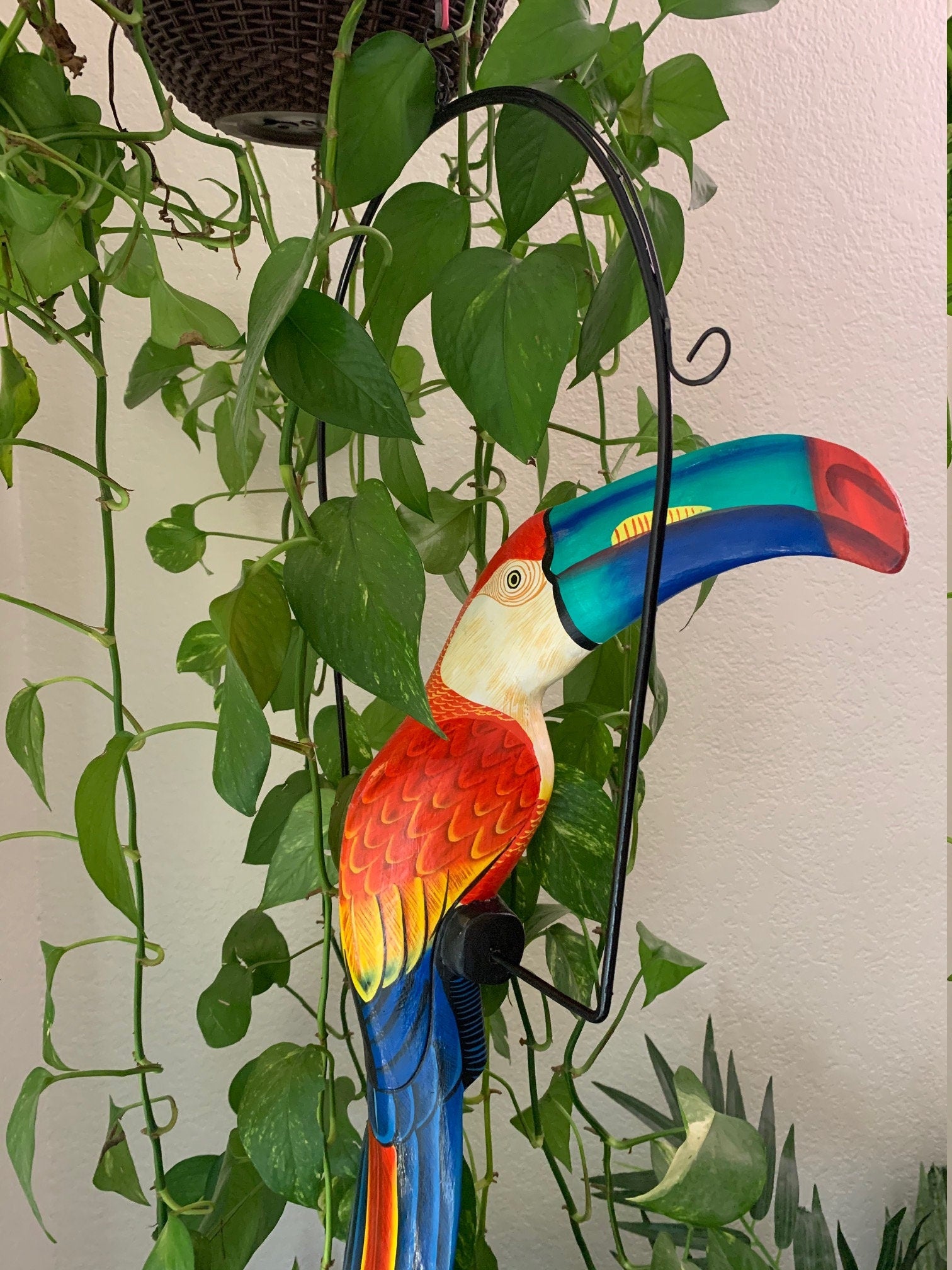 Colorful wooden parrot sculpture with a multicolored beak beside trailing green vines.