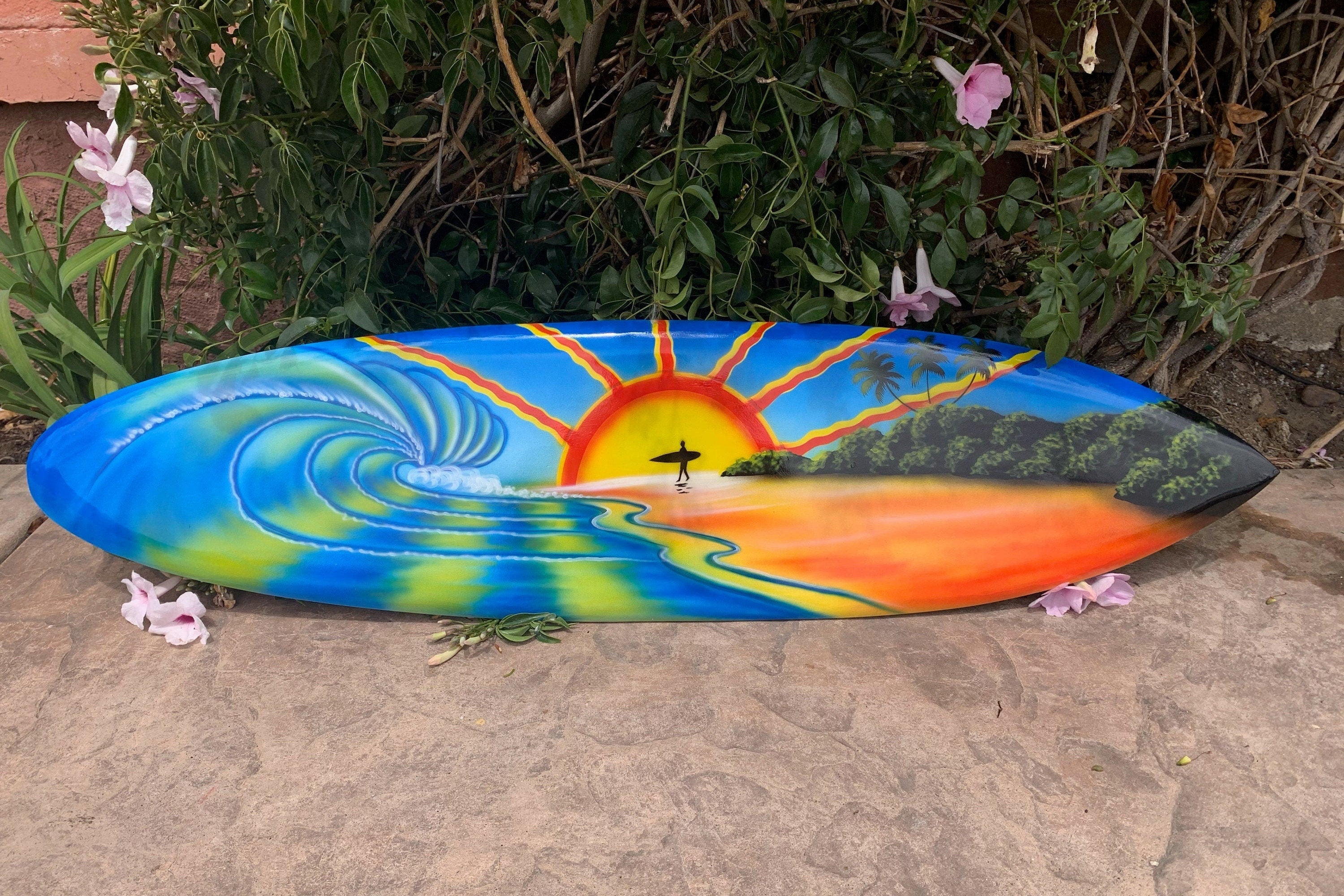 Colorful surfboard art depicting a sunset beach, waves, a surfer silhouette, and palm trees.