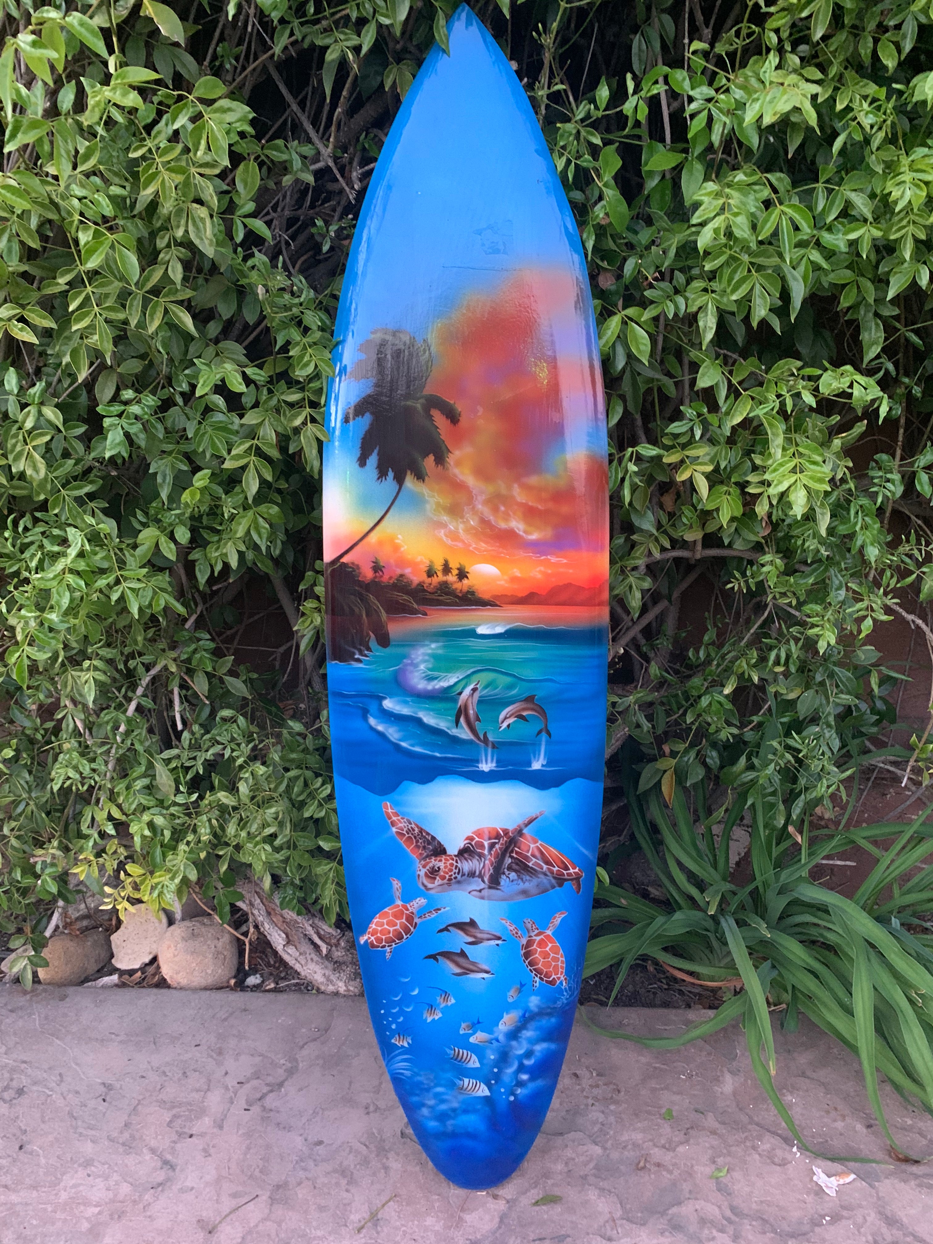 Colorful surfboard featuring tropical sunset, palm trees, dolphins and sea turtles against blue ocean.