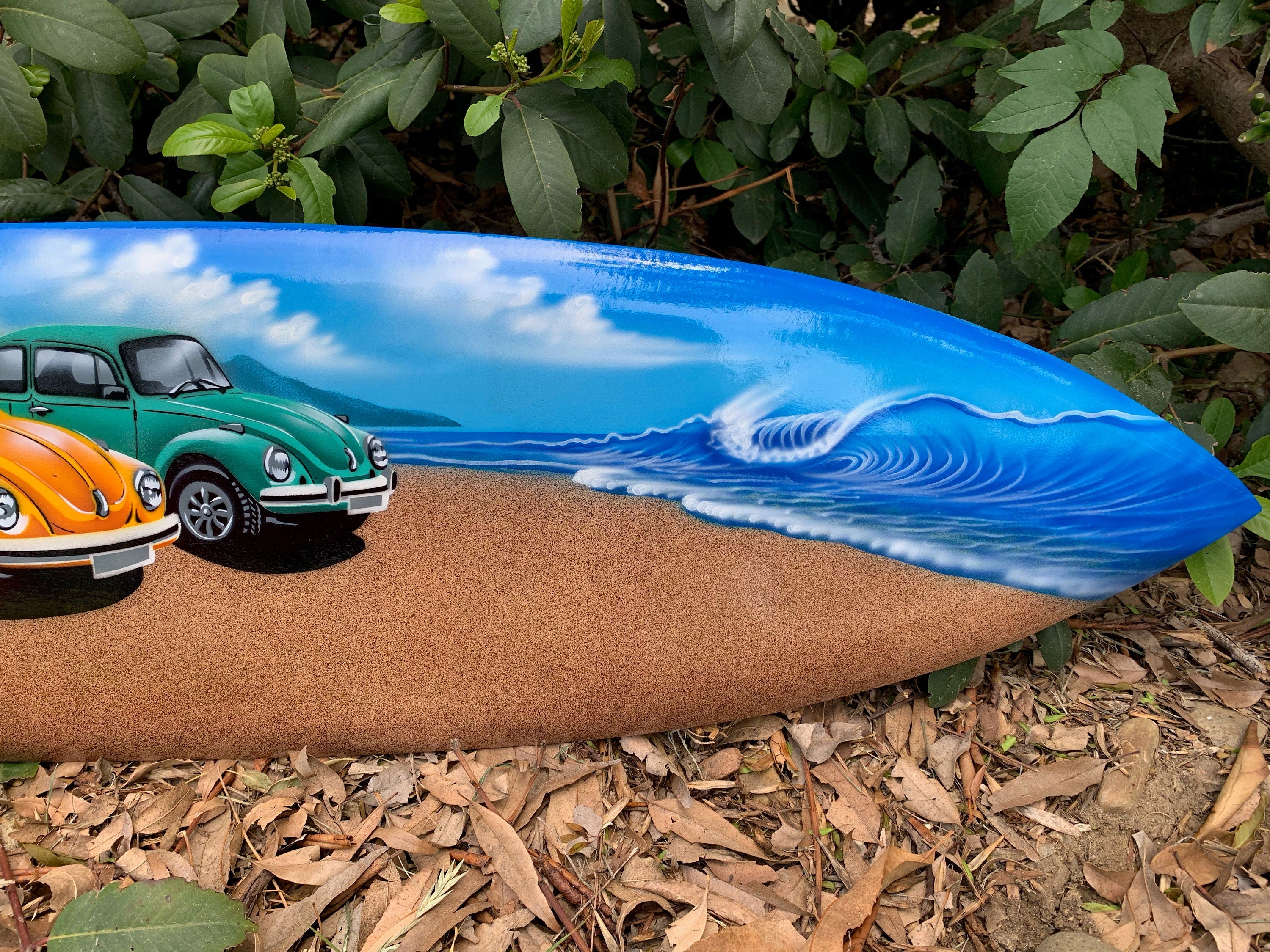 Colorful surfboard with vintage cars on a sandy beach and ocean waves.