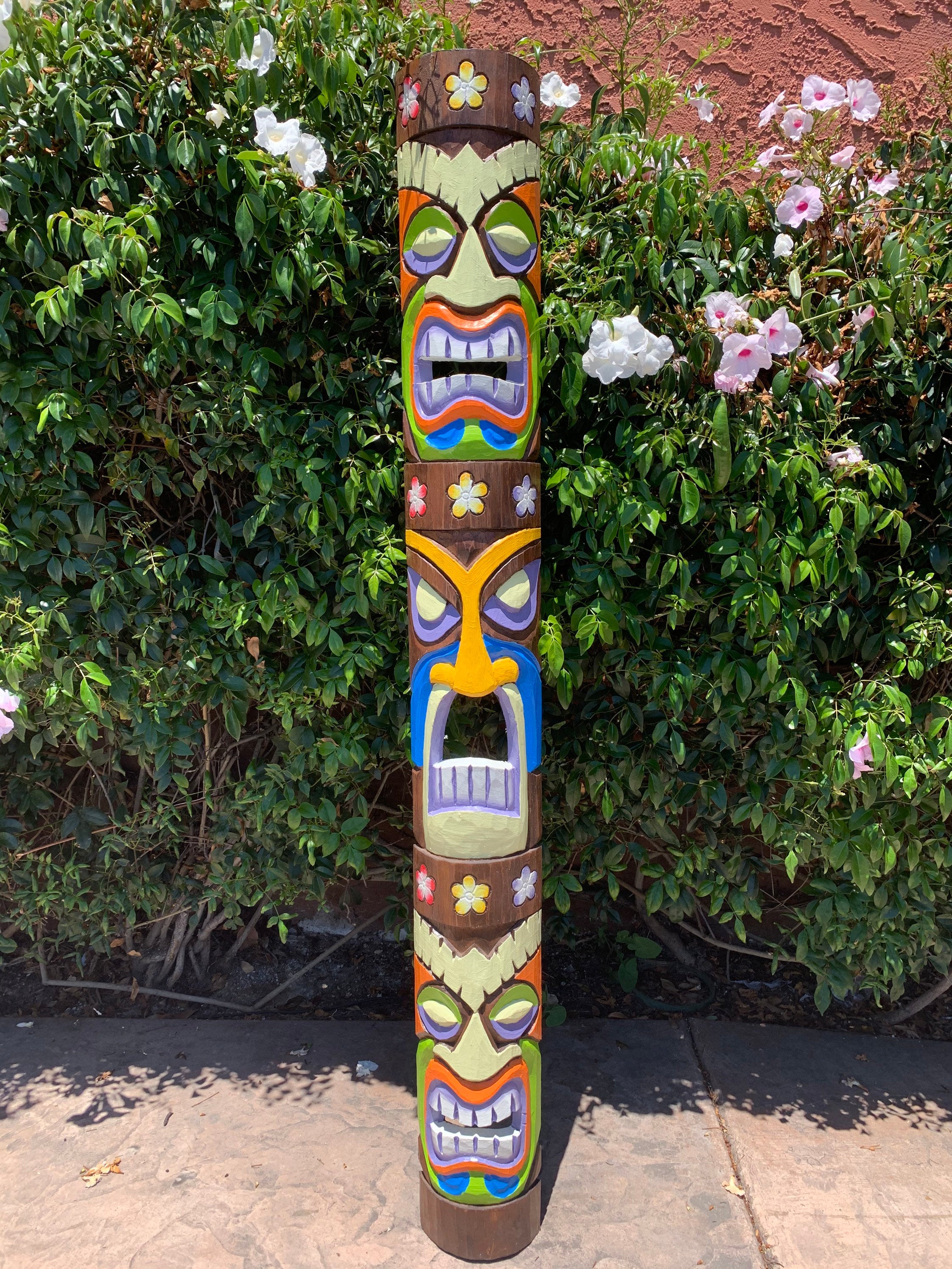 Colorful carved tiki totem pole outdoors among green bushes and white flowers.