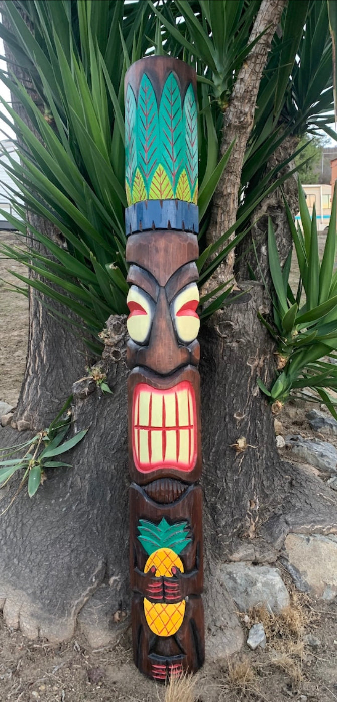 Colorful tiki totem pole with pineapple motif leaning against a tree in a garden.