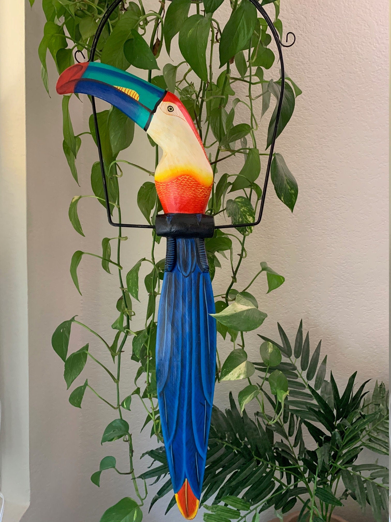 Colorful toucan sculpture with blue tail and bright beak, hanging among green houseplants.