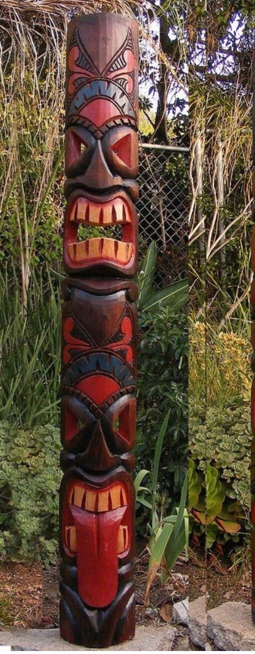 colorful wooden tiki totem pole statue in a garden setting with plants and a fence