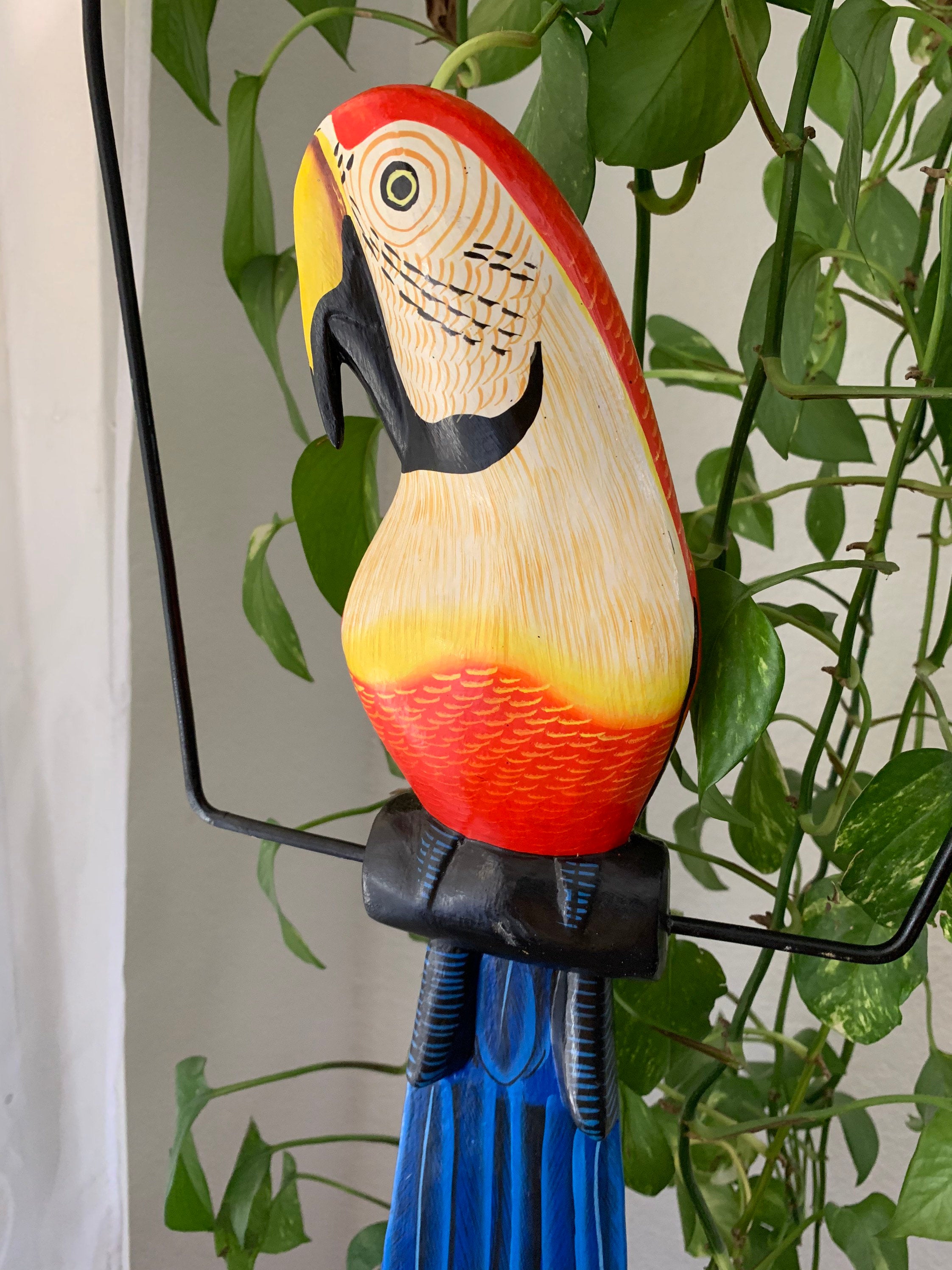 Colorful wooden toucan statue perched on a black stand with green leafy plants in the background.